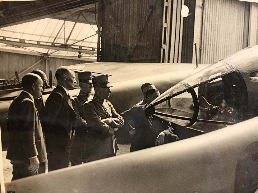 Sepia foto. Zes mannen, waarvan twee in uniform, bekijken de punt van een vliegtuig.