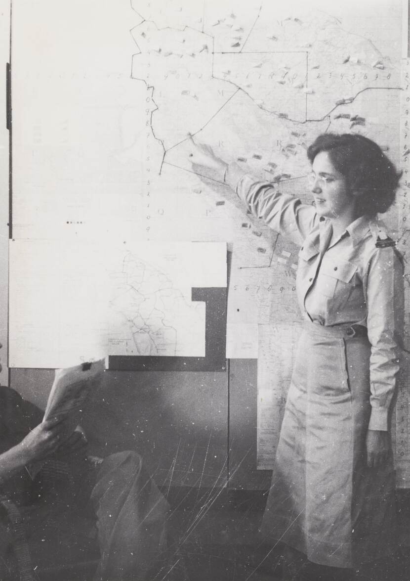 Zwart-witfoto, een vrouw in militair uniform wijst achter zich naar een kaart.