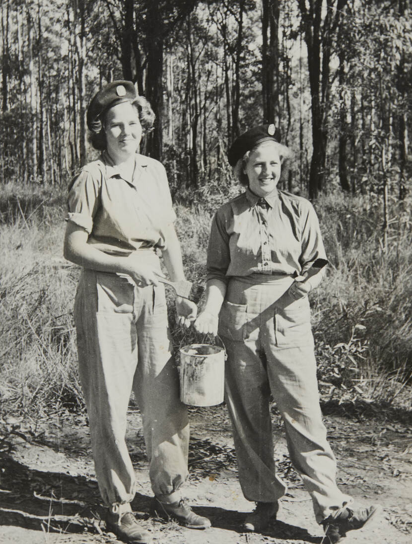 Zwart-witfoto van twee vrouwen in een overall, ze houden kwasten en een blik verf vast.