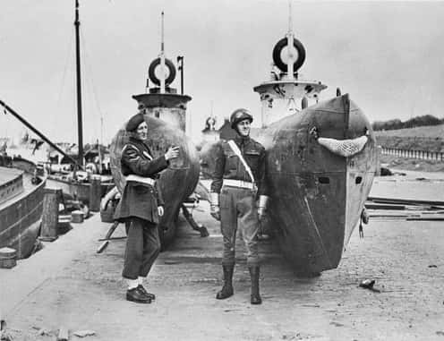 Zwart-witfoto, buiten. Twee militairen in uniform poseren bij twee aangemeerde Duitse onderzeeboten.