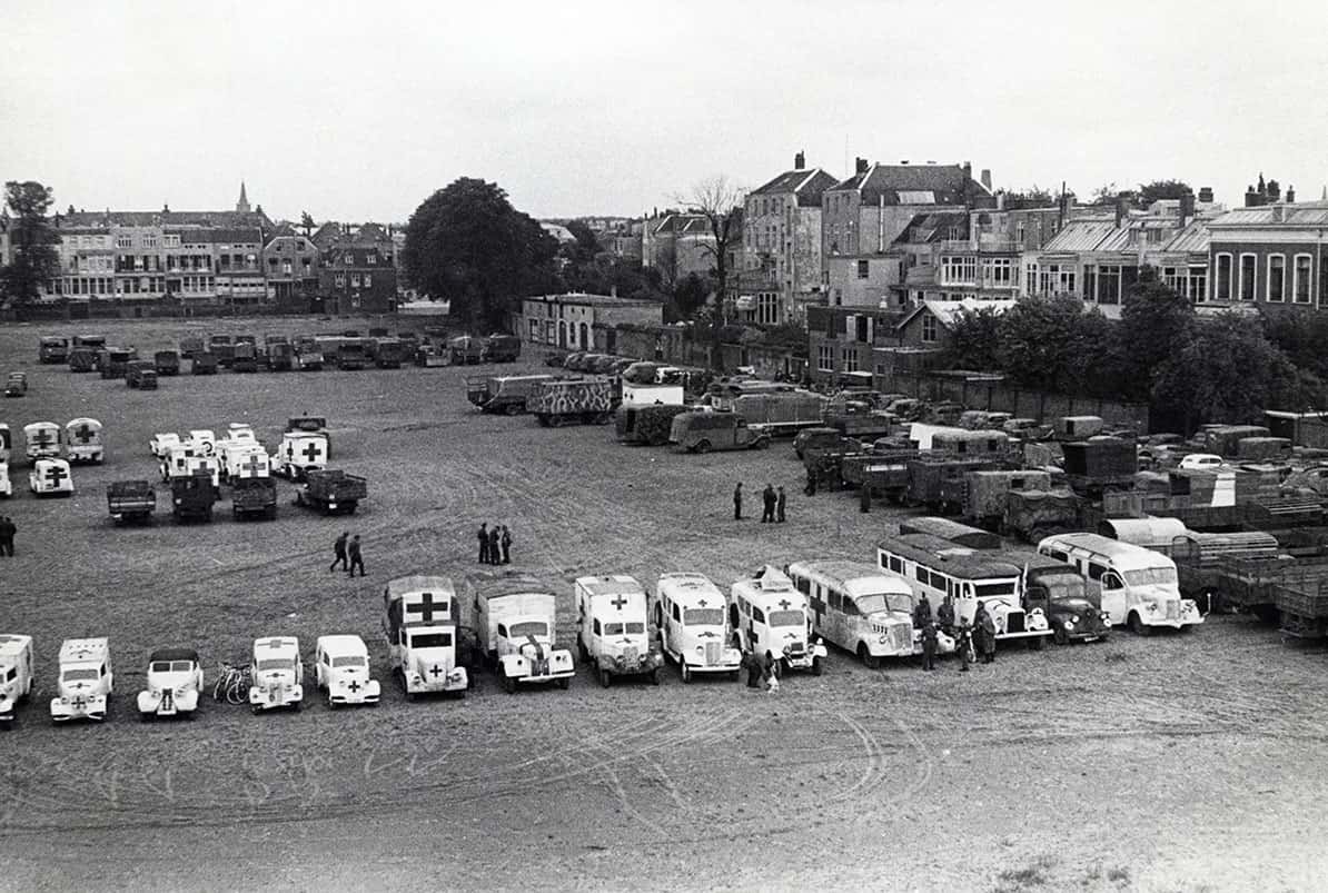 Zwart-witfoto, buiten. Een grasveld met daarop verschillende geparkeerde voertuigen.