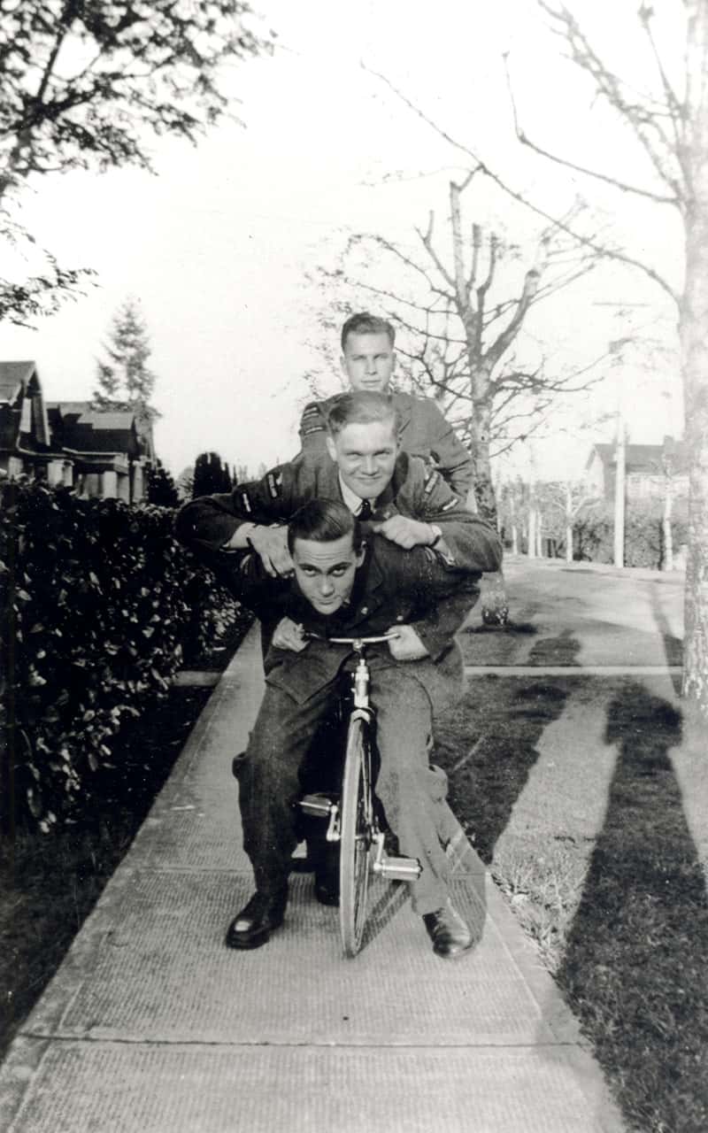 Zwart-witfoto. Drie militairen staan op de stoep. De voorste zit op een kleine fiets en leunt voorover over het stuur, de tweede hangt over hem heen. De derde staat er rechts achter. Ze dragen alle drie een net uniform en kijken in de camera. In de achtergrond een berm, bomen, links een heg en huizen. In de rechter benedenhoek is de schaduw van de fotograaf te zien.