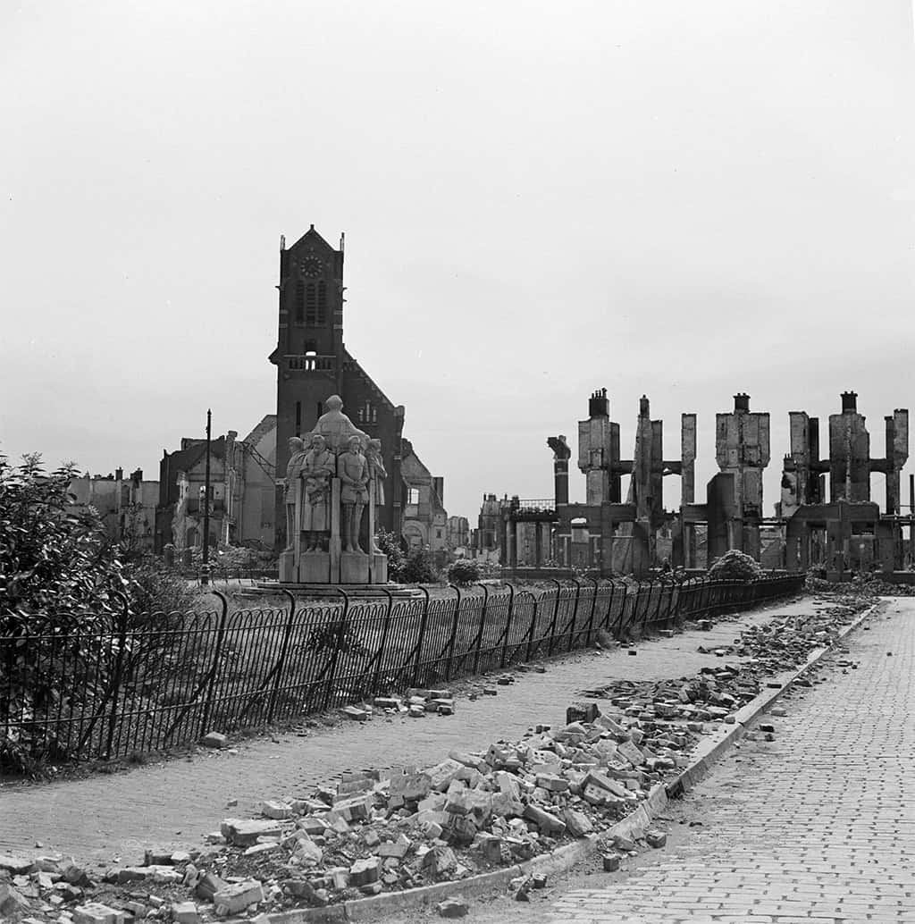 Zwart-witfoto. Een standbeeld in een park te midden van puin. Op de achtergrond staat een kerktoren.