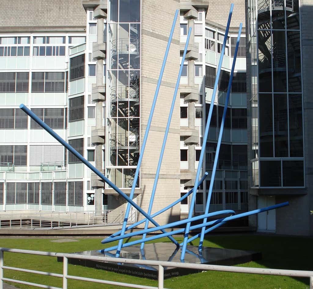 Kleurenfoto. Een constructie van blauwe palen op een grasveld voor flatgebouwen.