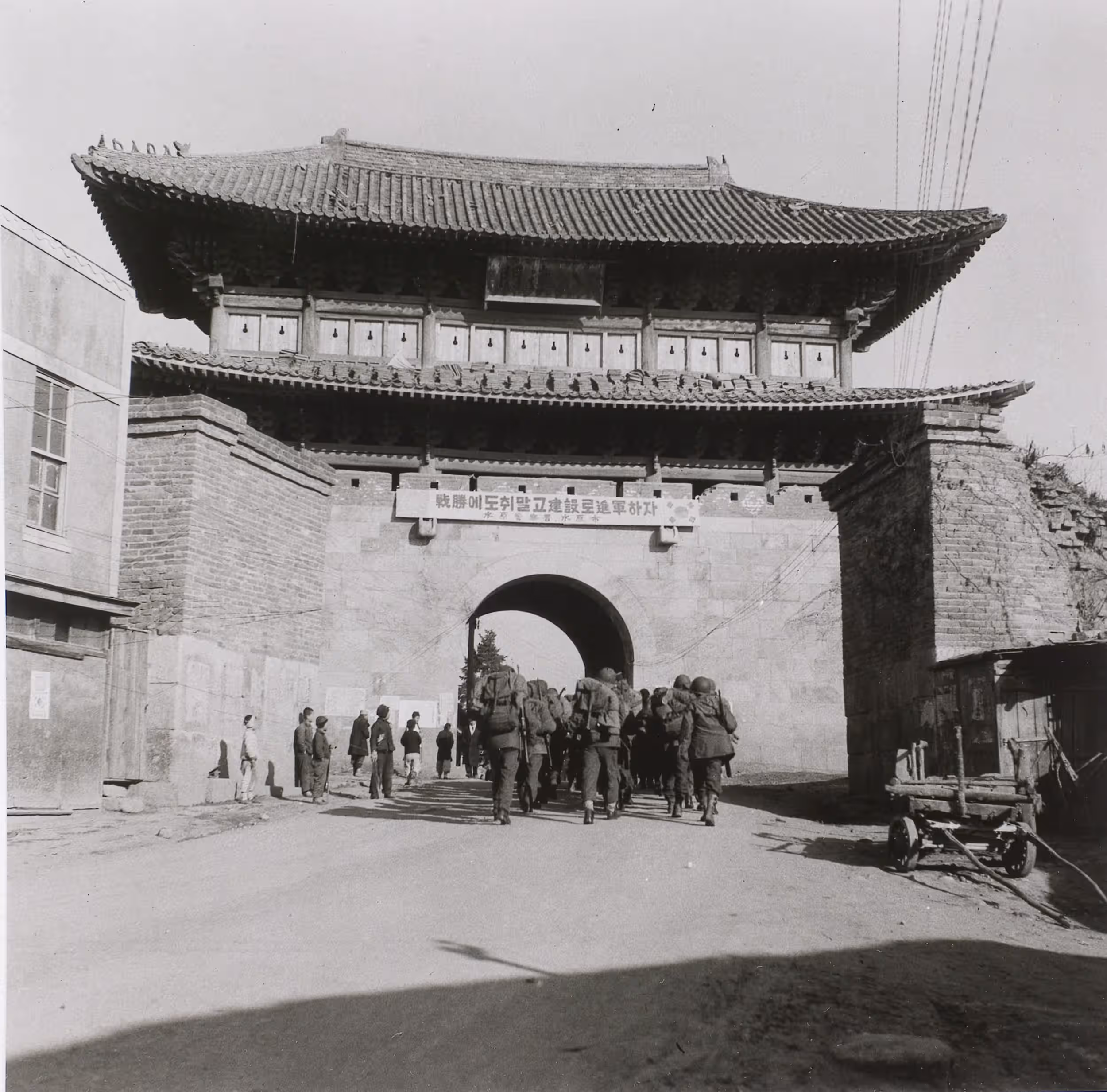 Zwart-witfoto, buiten op straat. Een grote, hoge poort met Oost-Aziatisch dak torent boven de straat uit, in het midden daarvan een doorgang. Over de weg loopt een groep zwaarbepakte militairen in rijen van drie door de doorgang. Links daarvan een groep mensen die staat toe te kijken, rechts een kar. Boven de doorgang een bord waar een Koreaanse tekst op staat geschreven.