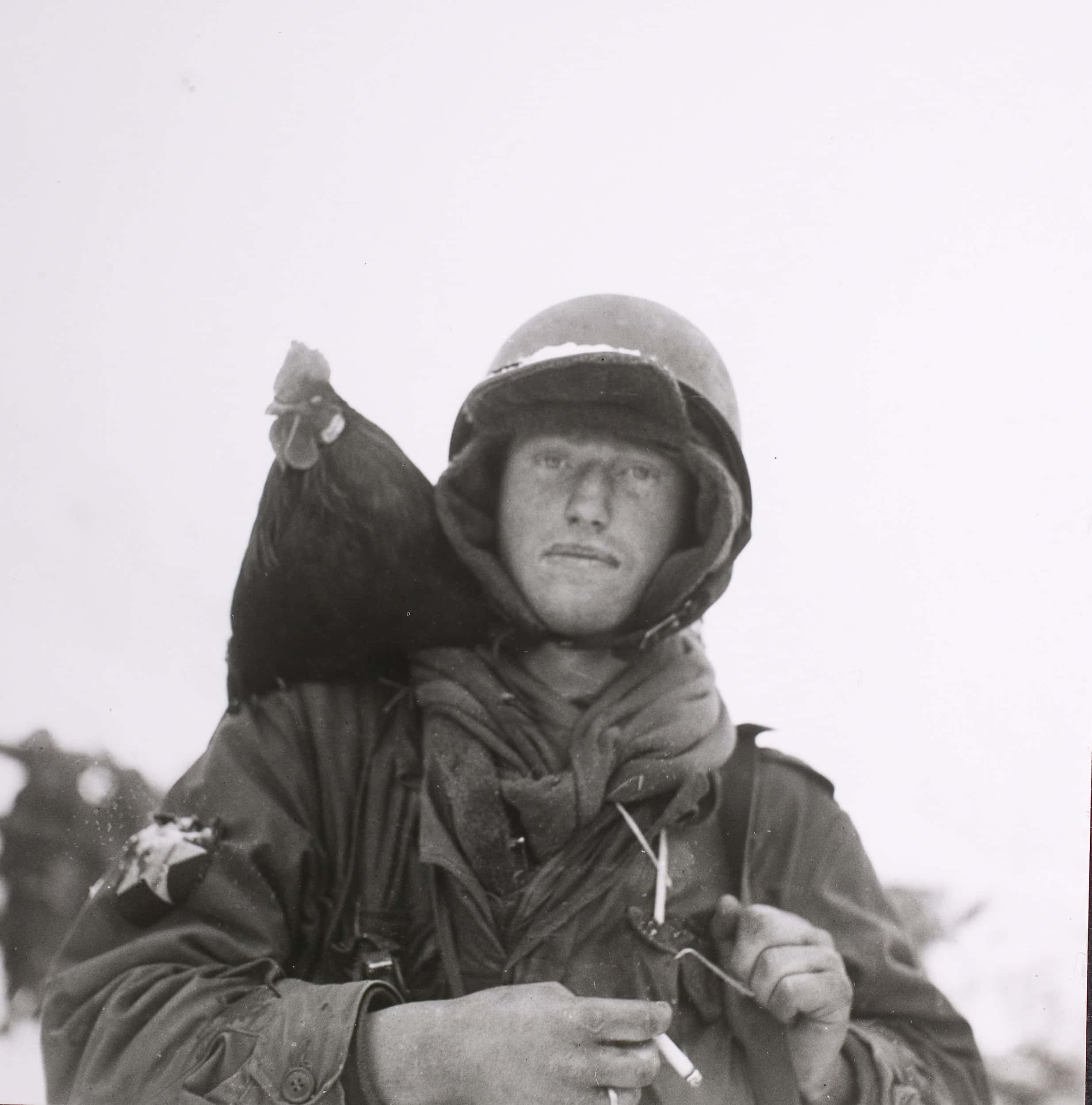 Zwart-witfoto, buiten. Een militair kijkt in de camera. Onder zijn helm draagt hij een gevoerde capuchon, een pet waarvan de klep onder de helm uitsteekt. Op de rand van zijn helm ligt een klein laagje sneeuw. Om zijn nek een sjaal. Tussen de vingers van zijn rechterhand heeft hij een sigaret, op zijn rechterschouder zit een kip.