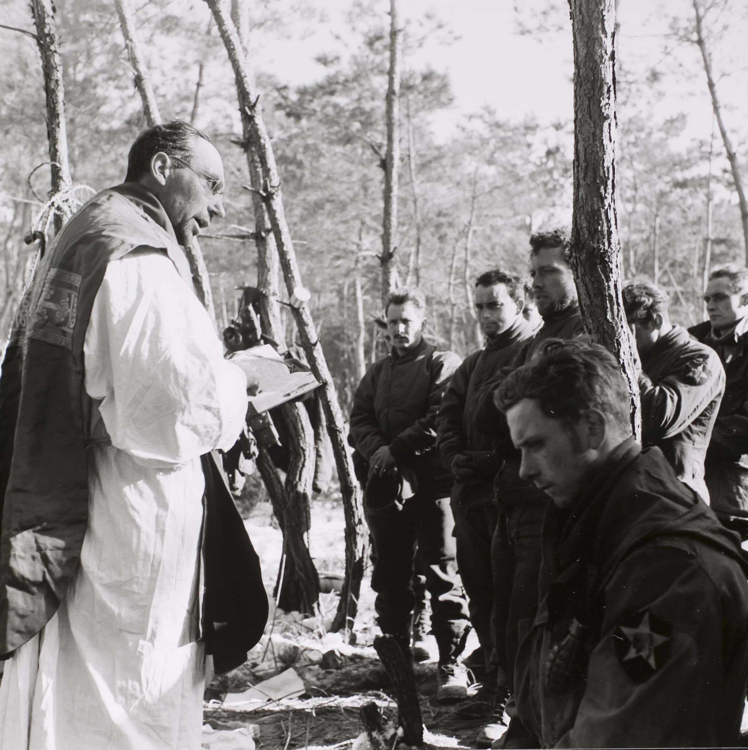 Zwart-witfoto, buiten in een bos. Links staat een man in priesterkleding, hij houdt een boek vast en leest daaruit voor, met zijn rechterwijsvinger op de pagina. Rechts een groep militairen die met serieuze gezichten luisteren, één van hen zit op z’n knieën en kijkt naar de grond, de anderen staan. Om hen heen bomen.