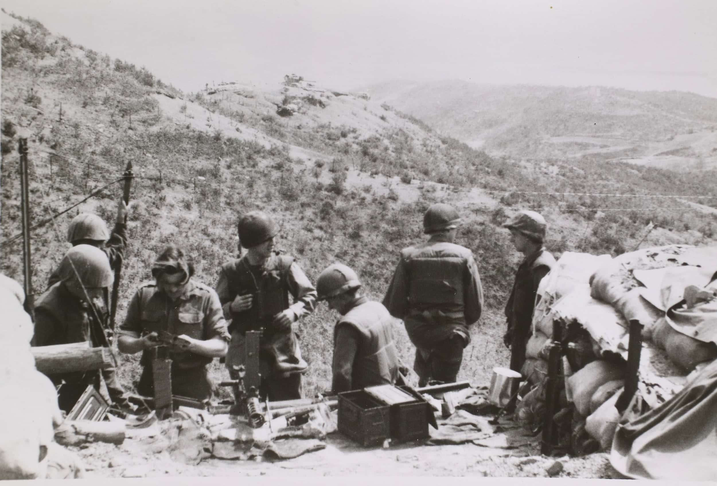 Zwart-witfoto, buiten in de bergen. Zeven militairen staan op een rij, voor hen liggen uit elkaar gehaalde wapens en munitiekisten, rechts een stapel zandzakken. Twee kijken naar de wapens, de rest kijkt om zich heen. Op de derde van links na, die een onderdeel van een wapen vasthoudt, draagt iedereen een helm. Op de achtergrond een bergachtig landschap, vooral bezaaid met lage struiken en jonge bomen.