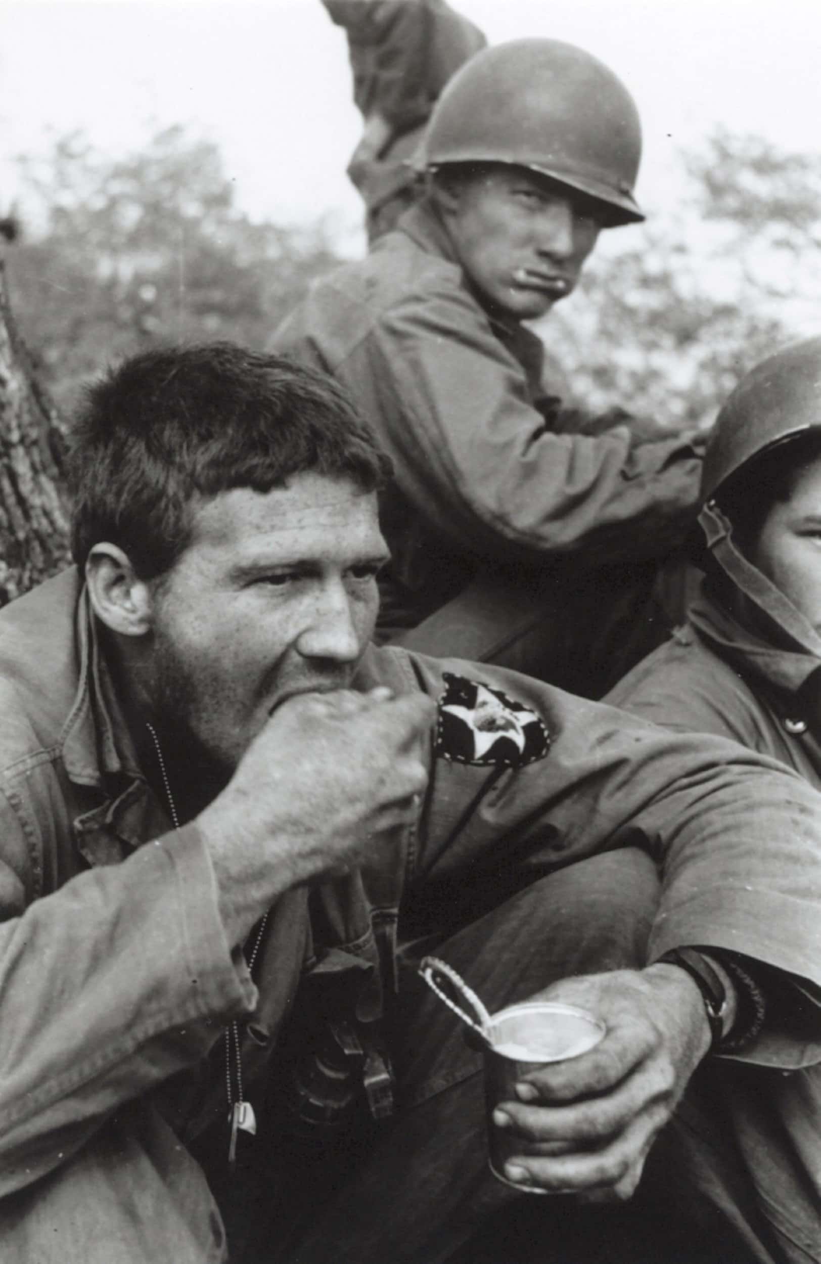 Zwart-witfoto, buiten. Een militair neemt een hap eten, terwijl hij met zijn linkerhand een opengemaakt blikje vasthoudt. Hij is blootshoofds, op zijn linkermouw een embleem van een donker schild met een witte ster. Achter hem kijkt een andere militair de camera in, hij draagt een helm en heeft zijn mond vol.