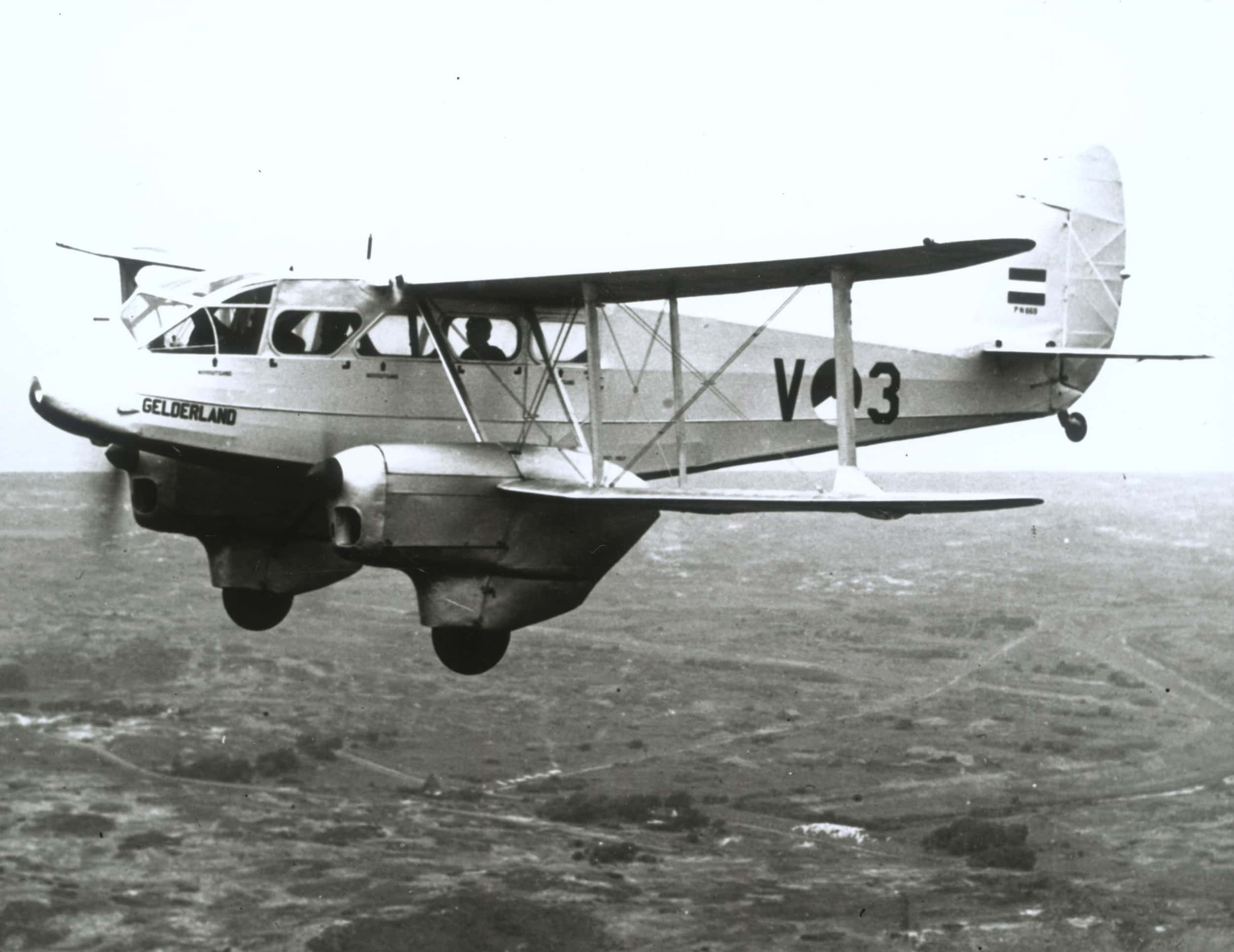 Zwart-witfoto, buiten in de lucht. Een wit vliegtuig vliegt door de lucht, op de zijkant bij de neus staat Gelderland, op de romp V3 met daartussen de rondel van de Koninklijke Luchtmacht. Op de staart is een kleine Nederlandse driekleur aangebracht.