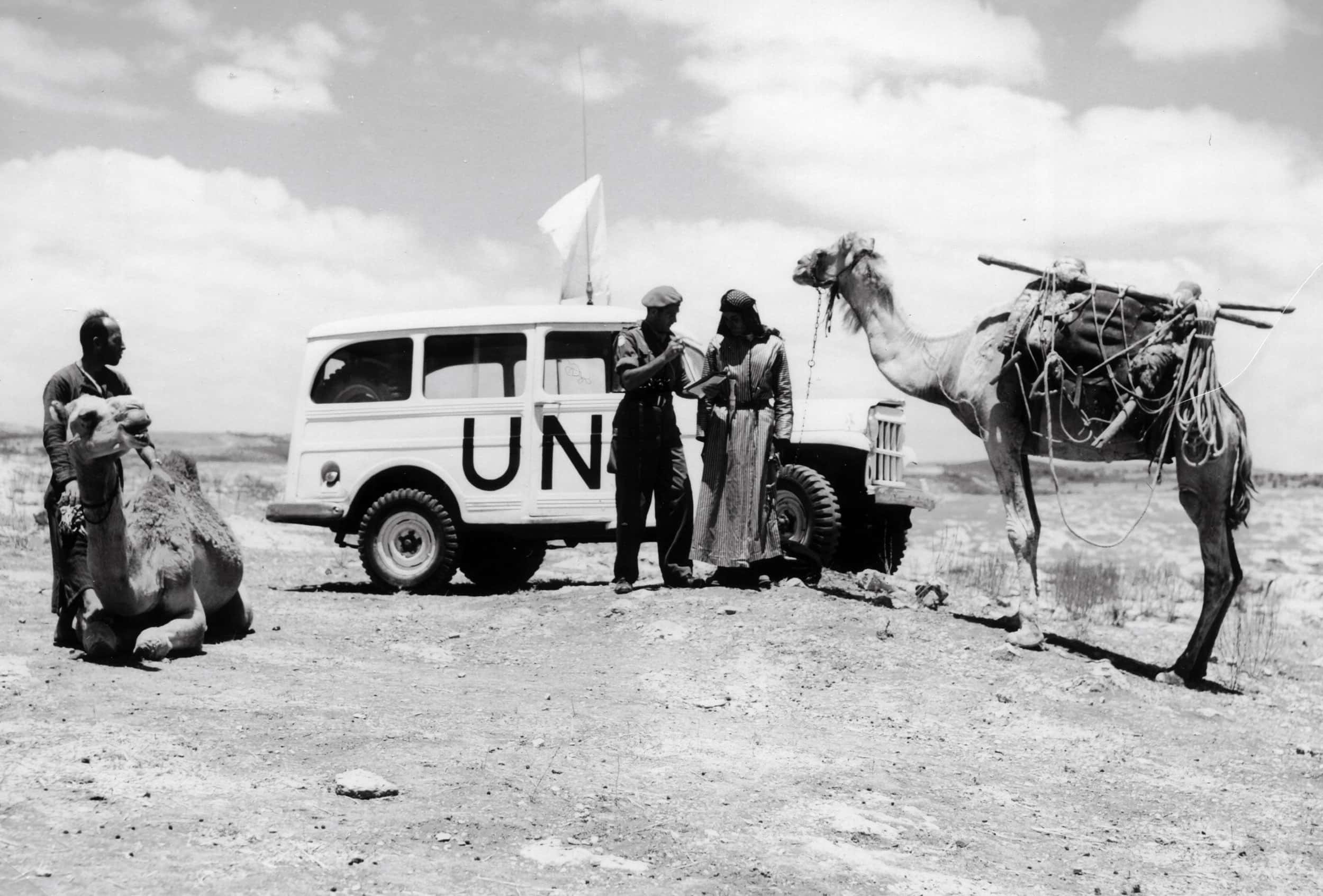 Zwart-witfoto, in de woestijn. Een witte jeep staat geparkeerd, op de zijkant staan de letters UN geschilderd en er wappert een witte vlag boven. Voor de auto staat een militair met een in gewaden geklede man te praten. Rechts van hen een grote dromedaris, voorzien van rijtuig, de man in gewaad houdt de teugels van het dier vast. Links staat een man met voor zich een andere dromedaris die op de grond ligt.