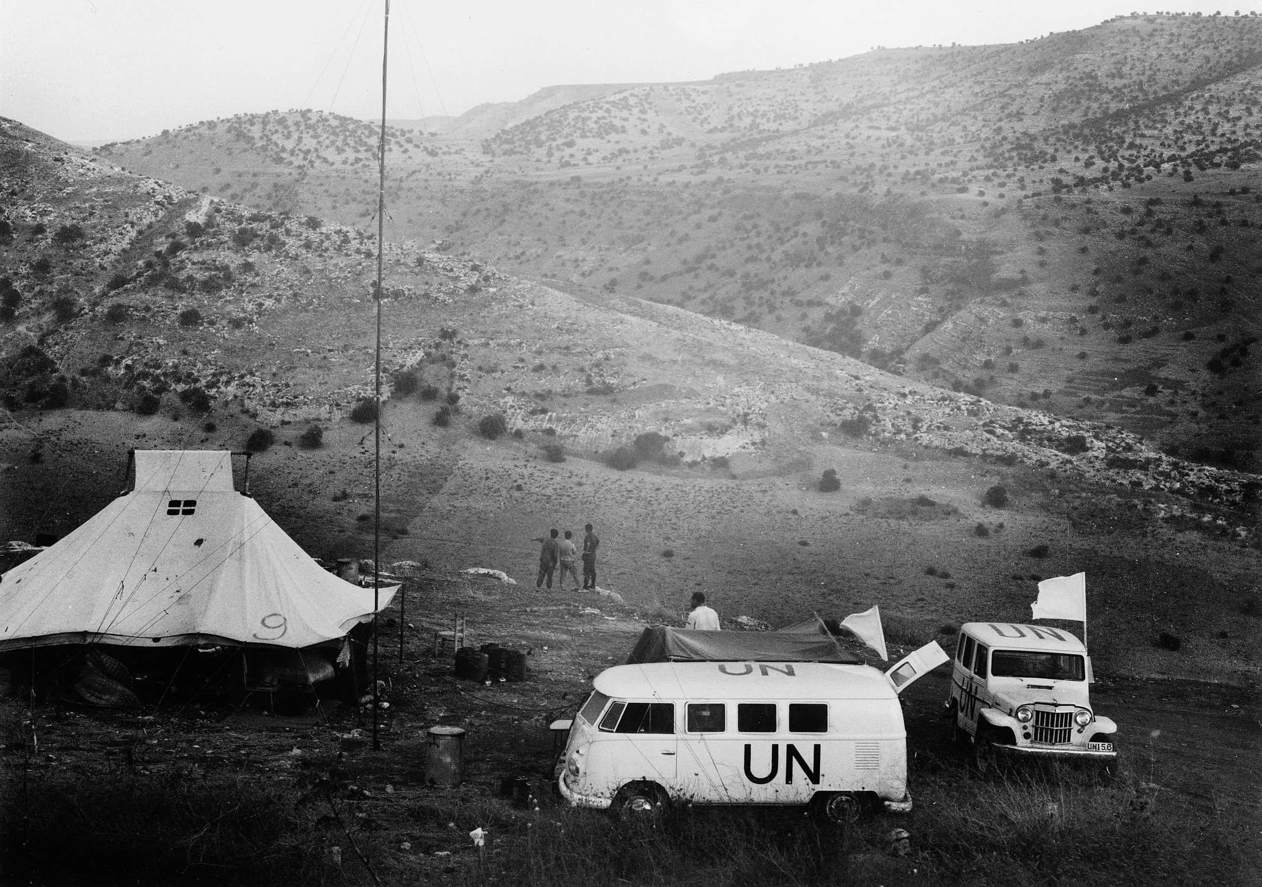 Zwart-witfoto, buiten in bergachtig gebied. Een busje en een jeep staan in een klein kampement geparkeerd, op beide voertuigen staan de letters UN geschilderd en beide hebben een vlaggenstok met witte vlag. Links een grote, witte tent, met het getal 9 erop. Tussen de tent en het busje een hoge zendmast, die met kabels aan de grond vaststaat. Drie mannen staan in een groepje bijeen, de linker wijst naar iets in de verte. Op de achtergrond een bergachtig landschap met lage struiken.