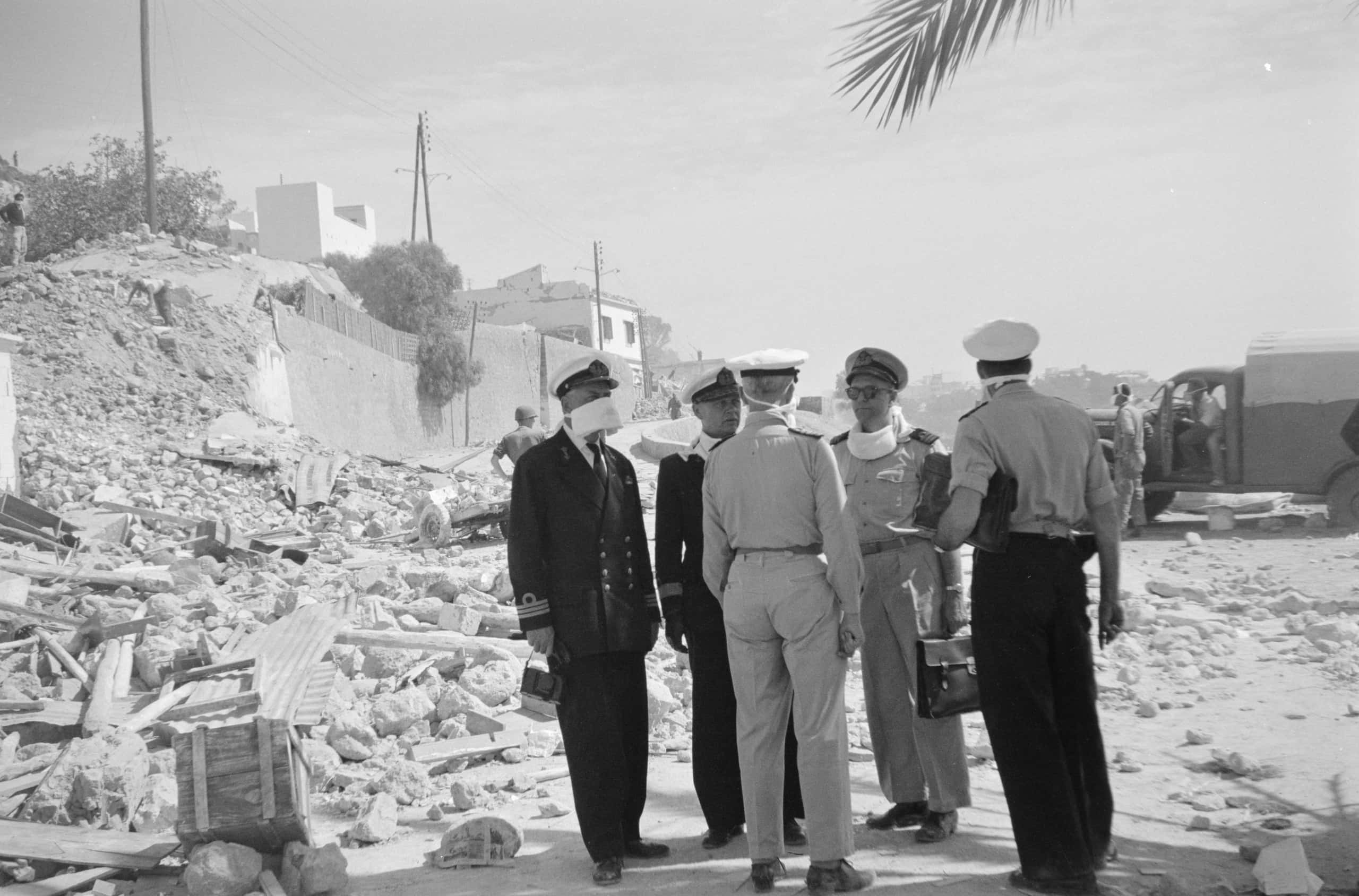 Zwart-witfoto, vijf marineofficieren, twee in donker pak, staan buiten in de schaduw van een boom. Links is puin van ingestorte gebouwen, rechts op de achtergrond een vrachtwagen.