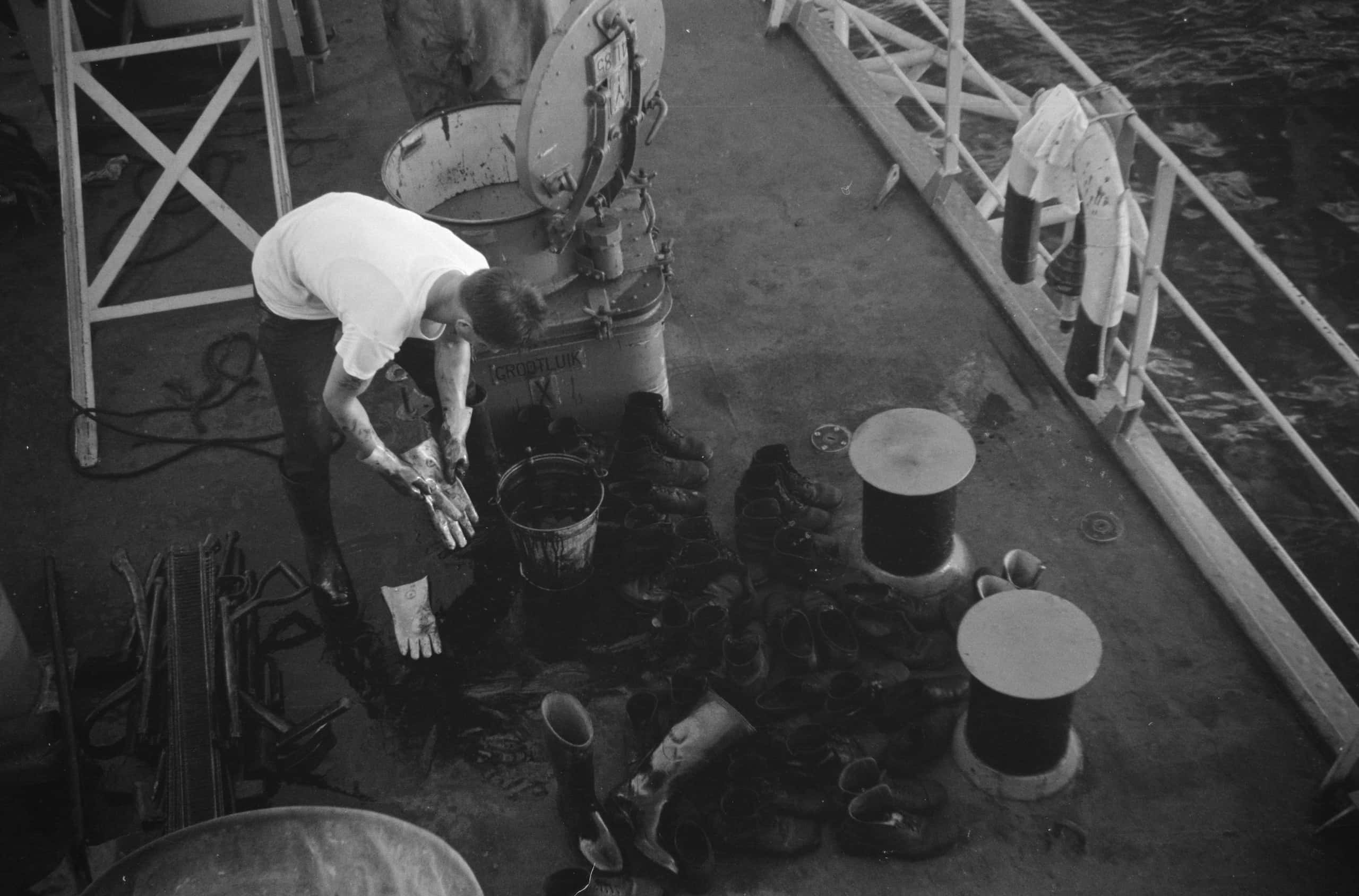 Zwart-witfoto, op het dek van een schip. Een man buigt voorover en wast een handschoen. Voor hem op de grond een emmer, laarzen en schoenen. Naast hem ligt een stapel scheppen.