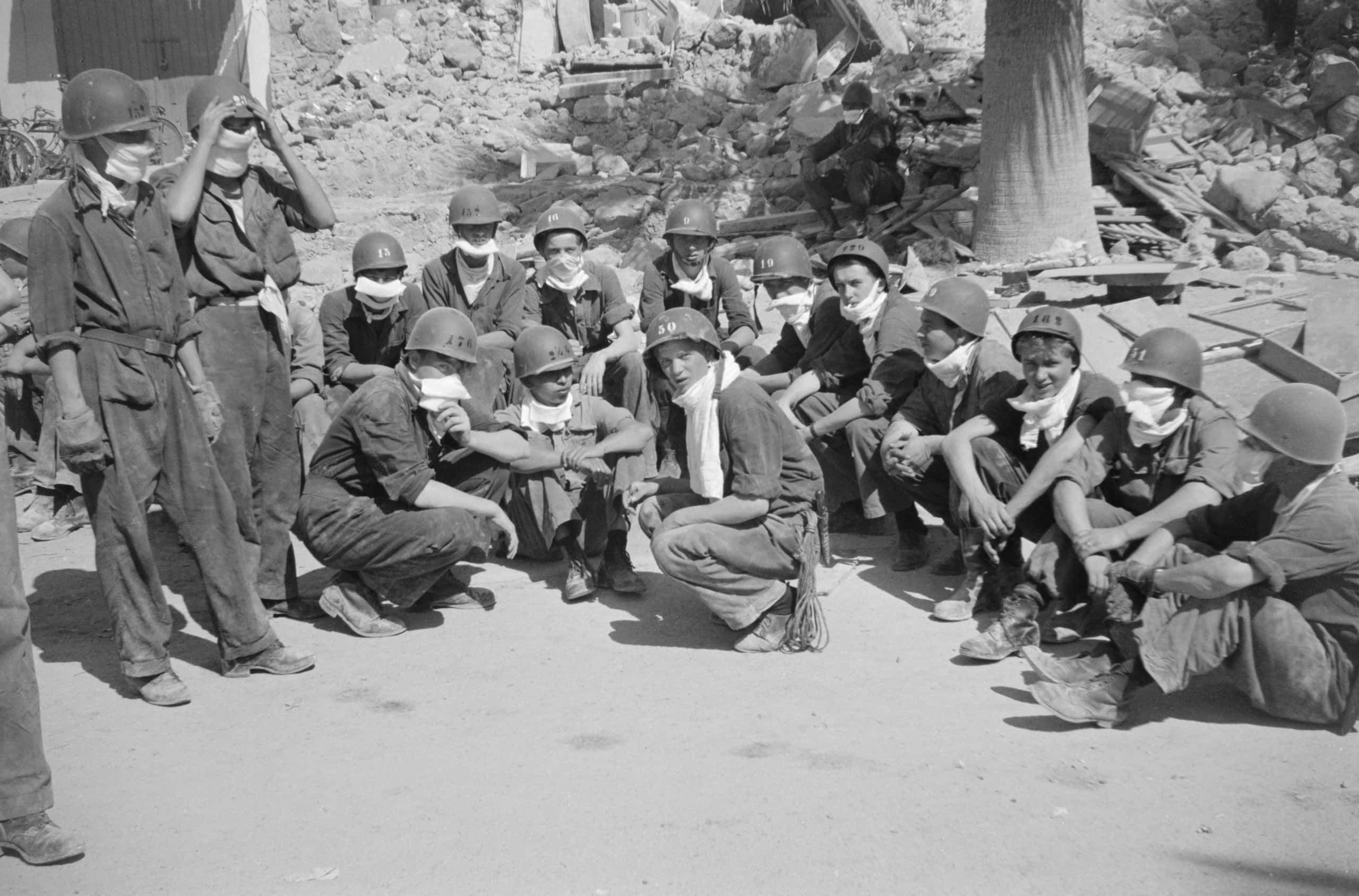 Zwart-witfoto, vijftien militairen met helmen op zitten op de grond. Twee helemaal links staan, twee anderen zitten op de hun hurken. Sommigen dragen een masker voor de mond. Op de achtergrond de voet van een boom en puin waar een oudere man op zit.