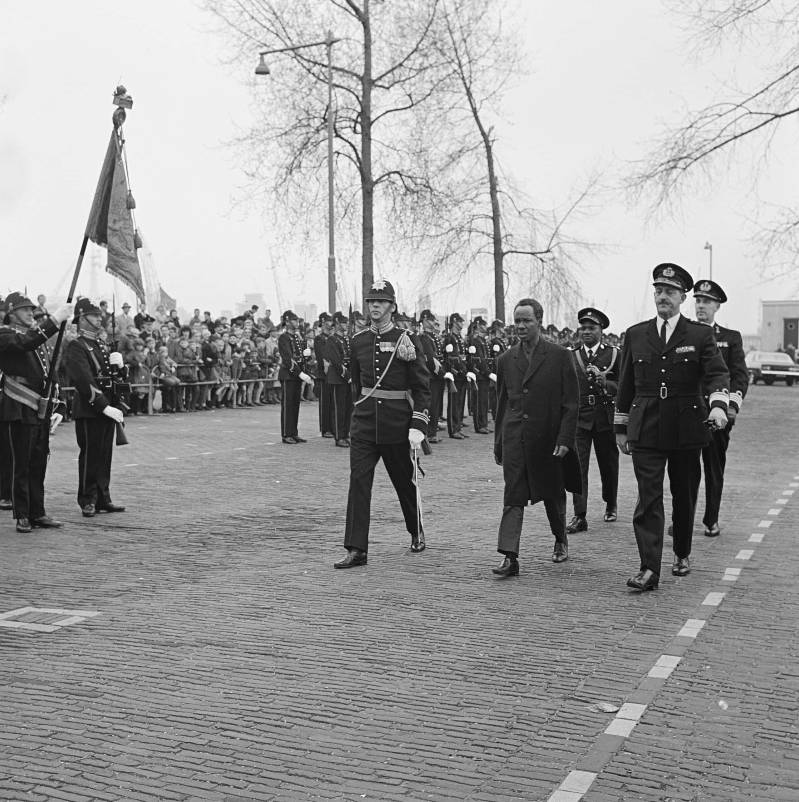 Zwart-witfoto, vijf mannen lopen over een parkeerplaats, drie voor, twee daarachter. De man middenvoor draagt een lange zwarte jas, de rest is in militair uniform. Links een vaandelwacht die een vaandel omhoog houdt, daarnaast, half achter het lopende groepje, een opgestelde erewacht in de houding. Daar weer achter staat publiek achter dranghekken toe te kijken. Op de achtergrond enkele bomen en een lantaarnpaal.