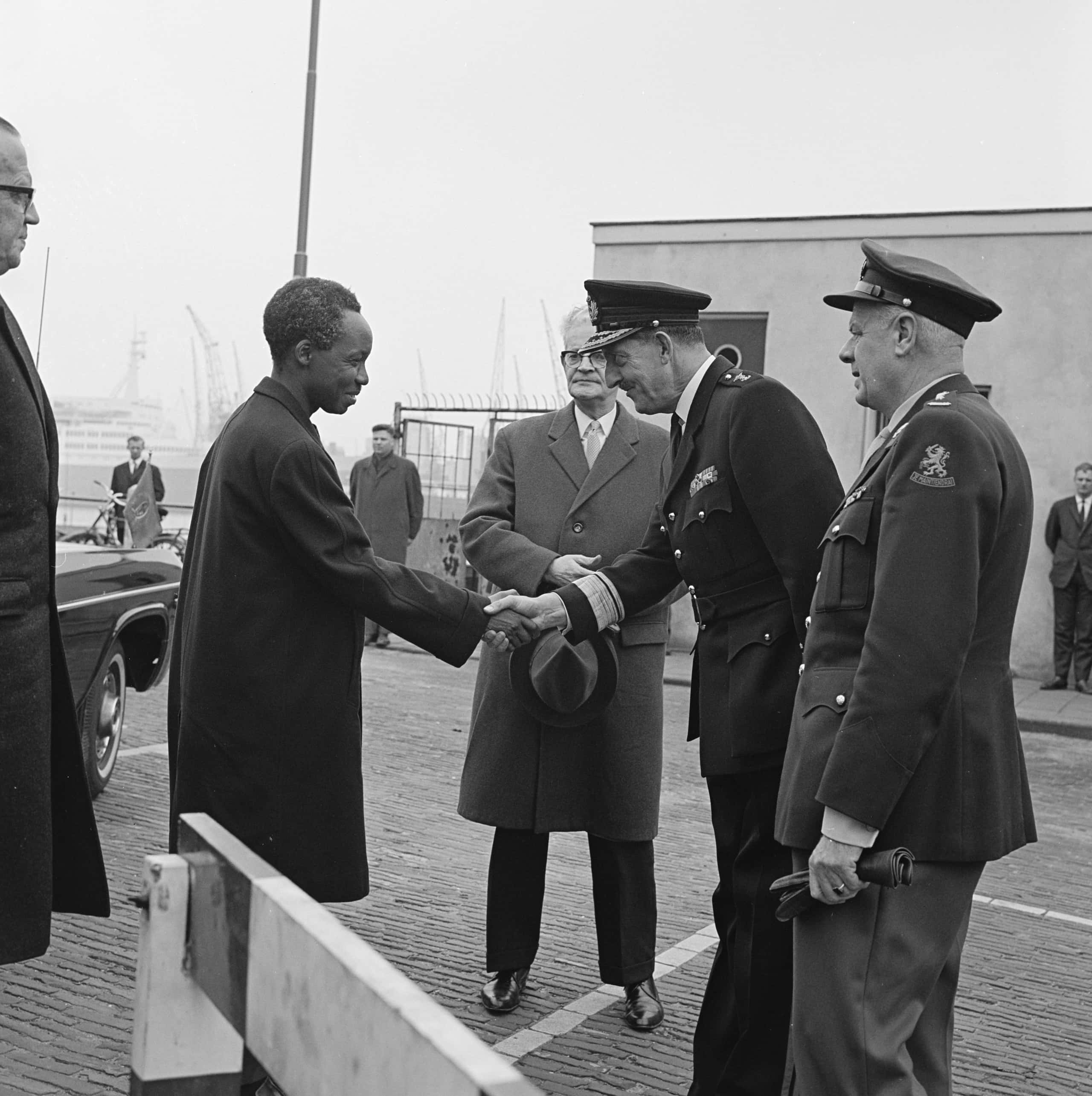 Zwart-witfoto, twee mannen schudden elkaar de hand, onder toeziend oog van drie anderen. De man links draagt een lange zwarte jas, de man rechts het uniform van een generaal, voorzien van strepen, sterren en onderscheidingen. Links is een klein hoekje van een auto te zien, op de achtergrond een groot schip en hijskranen van een haven.