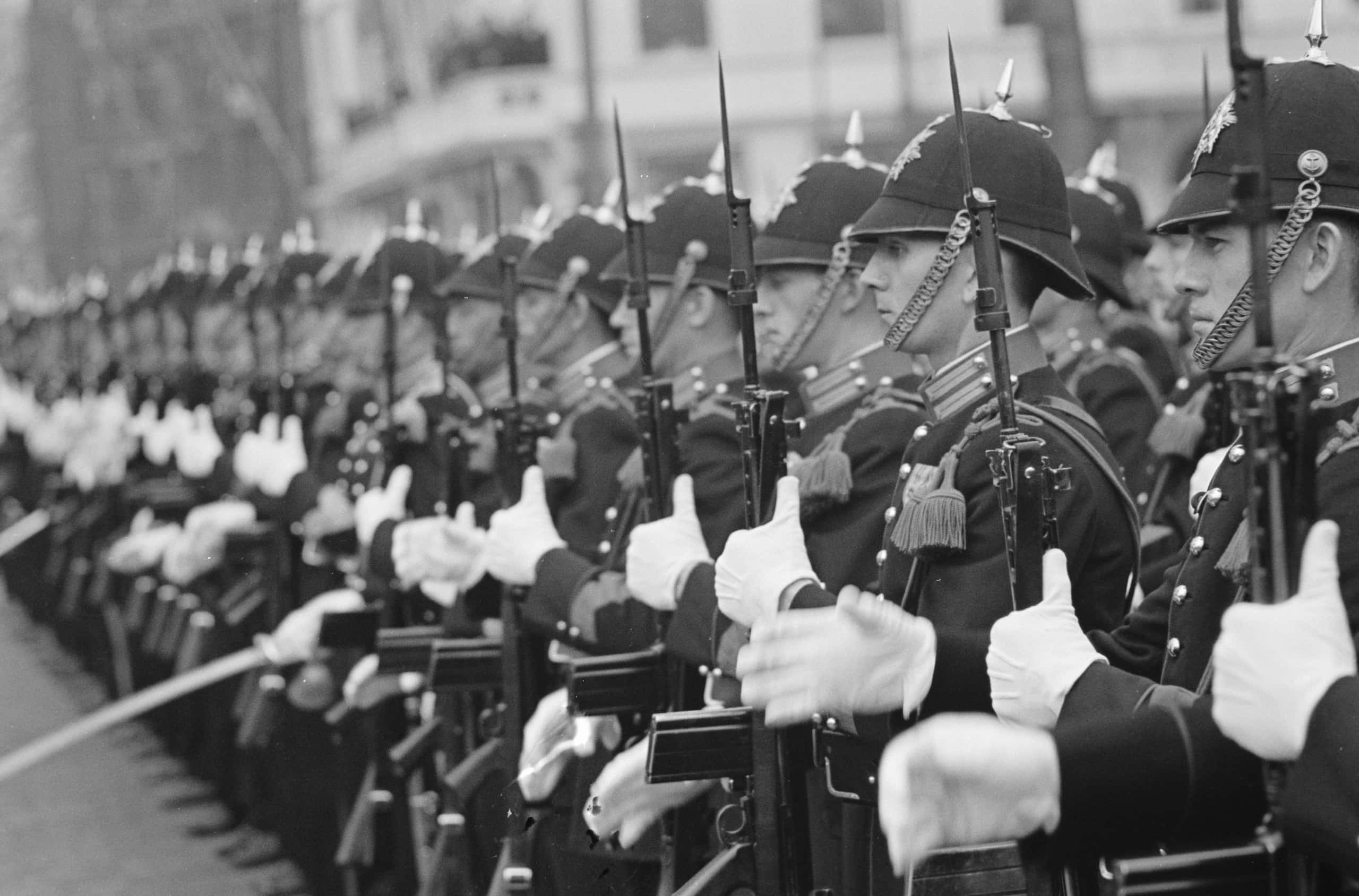 Zwart-witfoto, tientallen militairen staan in een rij opgesteld, zij dragen ceremoniële uniformen met koorden, kwarten en witte handschoenen, op hun hoofd een pikethelm die met een riempje om hun kin zit. Zij houden een geweer recht overeind voor hun borst. De scherpheid van de foto naar links toe steeds meer af.