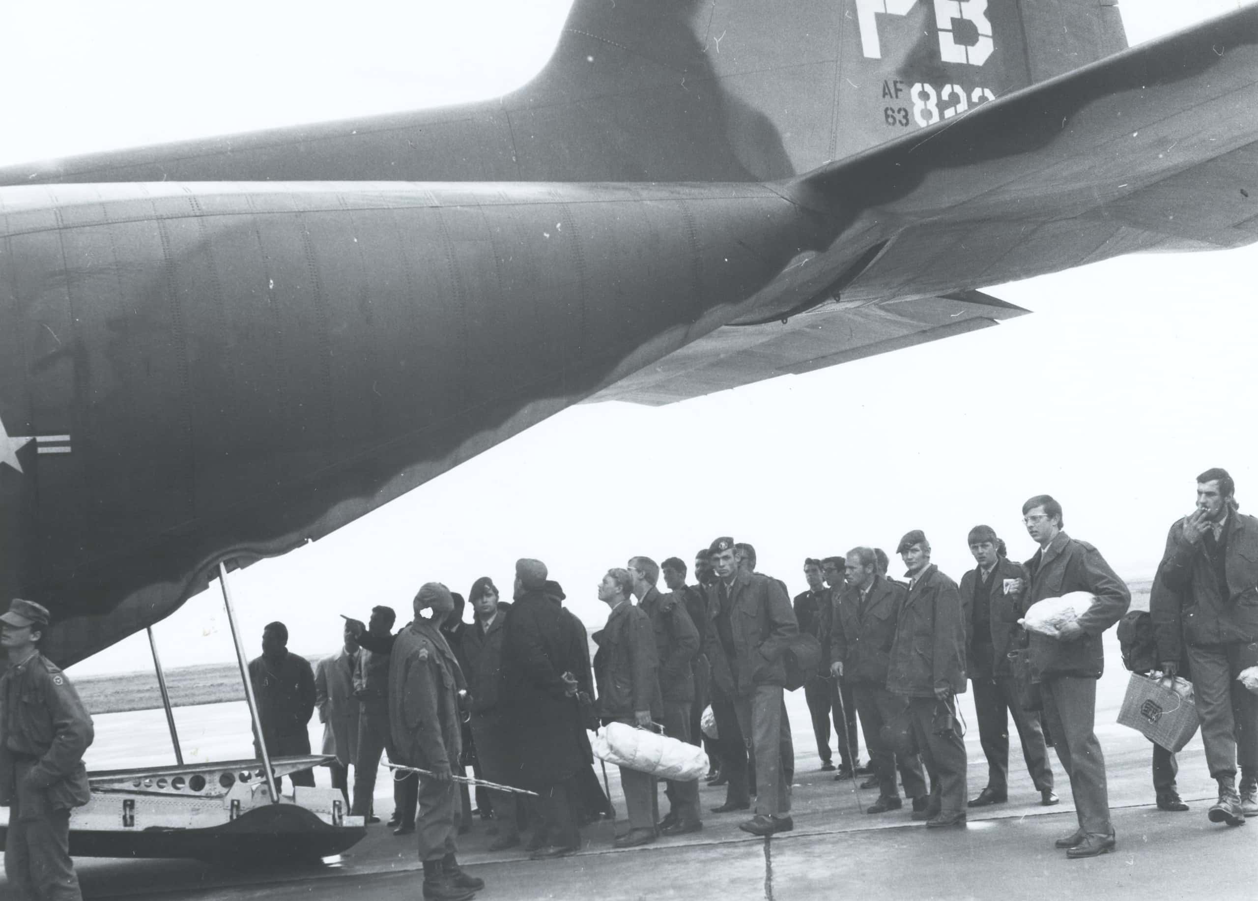 Zwart-witfoto, een grote groep mannen in legerkleding staat in een rij te wachten. Links en boven hen steekt de achterkant van een groot vliegtuig uit.