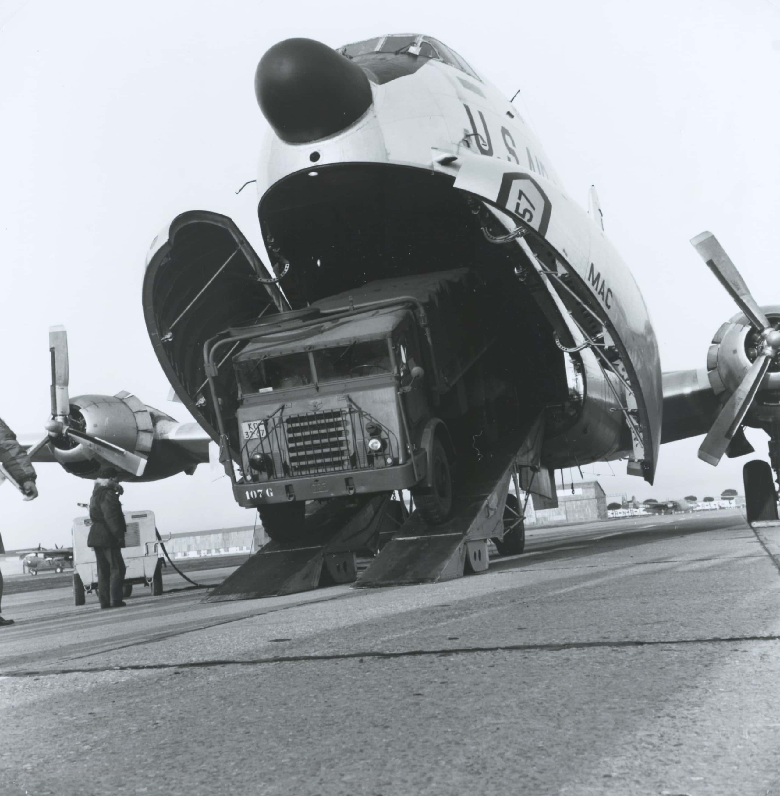 Zwart-witfoto, een vliegtuig van voren gezien die vanaf de onderkant open is. Uit het vliegtuig komt een vrachtwagen gereden. Links staat een man op de vrachtwagen te wachten.