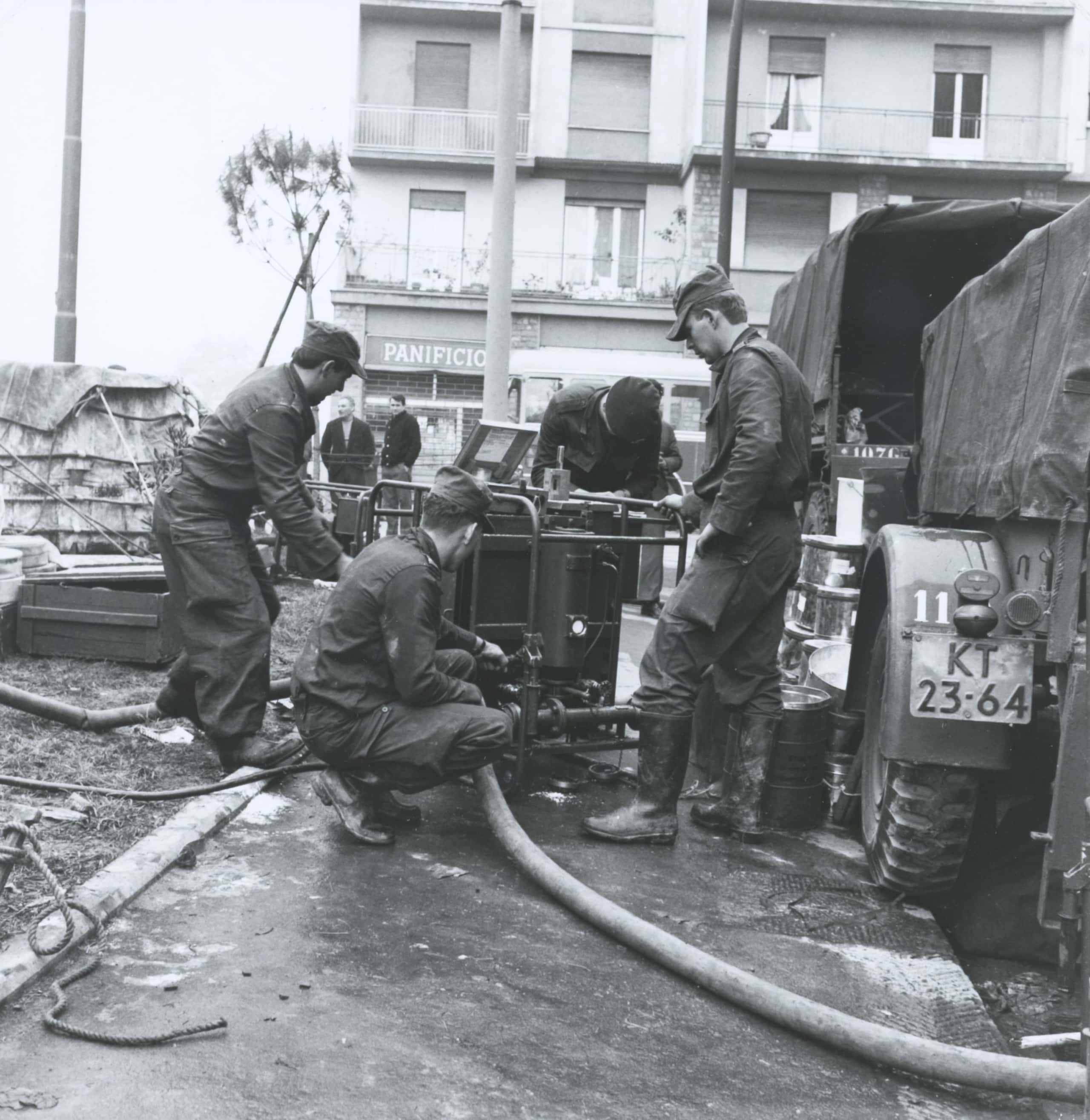 Zwart-witfoto, vier mannen in legerkleding werken aan een waterpomp. Op de grond liggen meerdere waterslangen. Op de achtergrond kijken twee andere mannen toe, rechts staat een vrachtwagen met aanhanger, van achteren gezien.