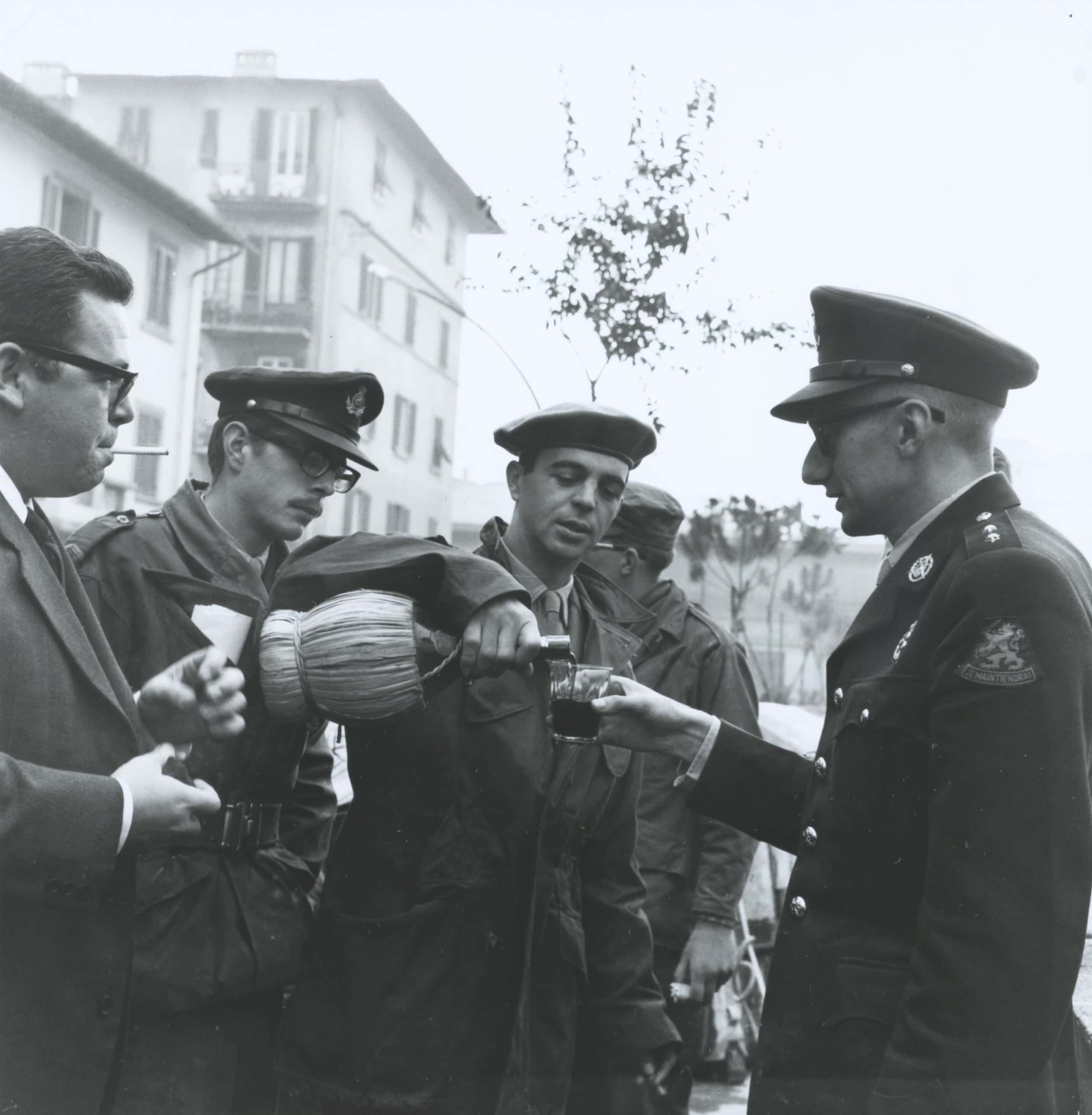 Zwart-witfoto, vijf mannen staan op straat. Vier dragen legerkleding. De man rechts houdt een glas vast en krijgt van de man in het midden wijn ingeschonken. De man helemaal links rookt een sigaret.