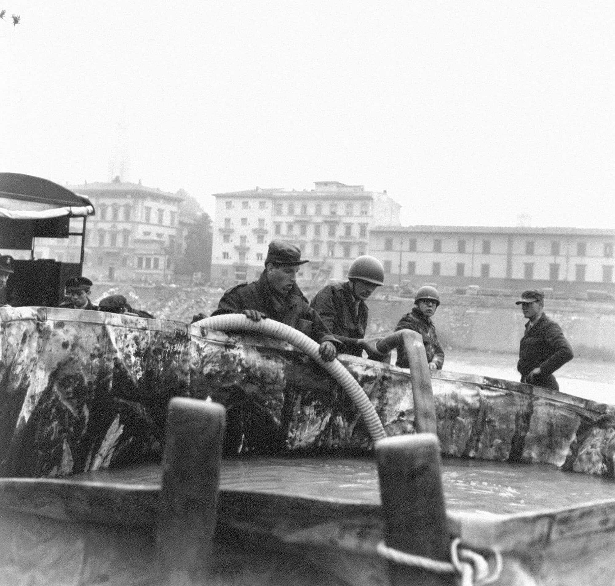Twee mannen in legerkleding houden waterslangen in een grote bak met water. Om hen heen zijn nog vijf anderen ook aan het werk. Ze staan op de oever van een rivier, die op de achtergrond te zien is.