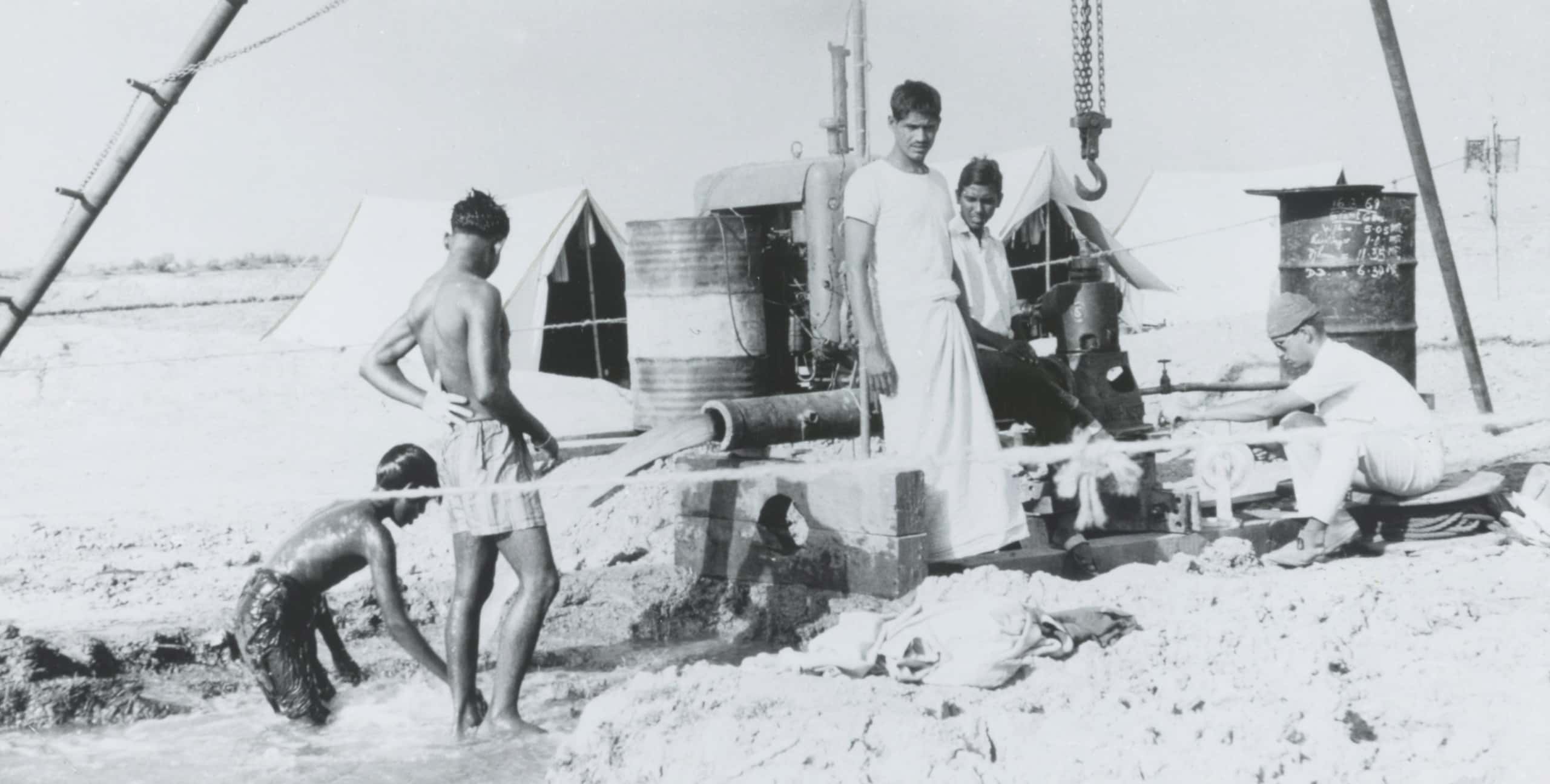 Zwart-witfoto, een groep mannen staan om een buis waar water uit komt. Op de achtergrond tenten en olievaten. Boven de pomp aan de buis hangt een grote haak.