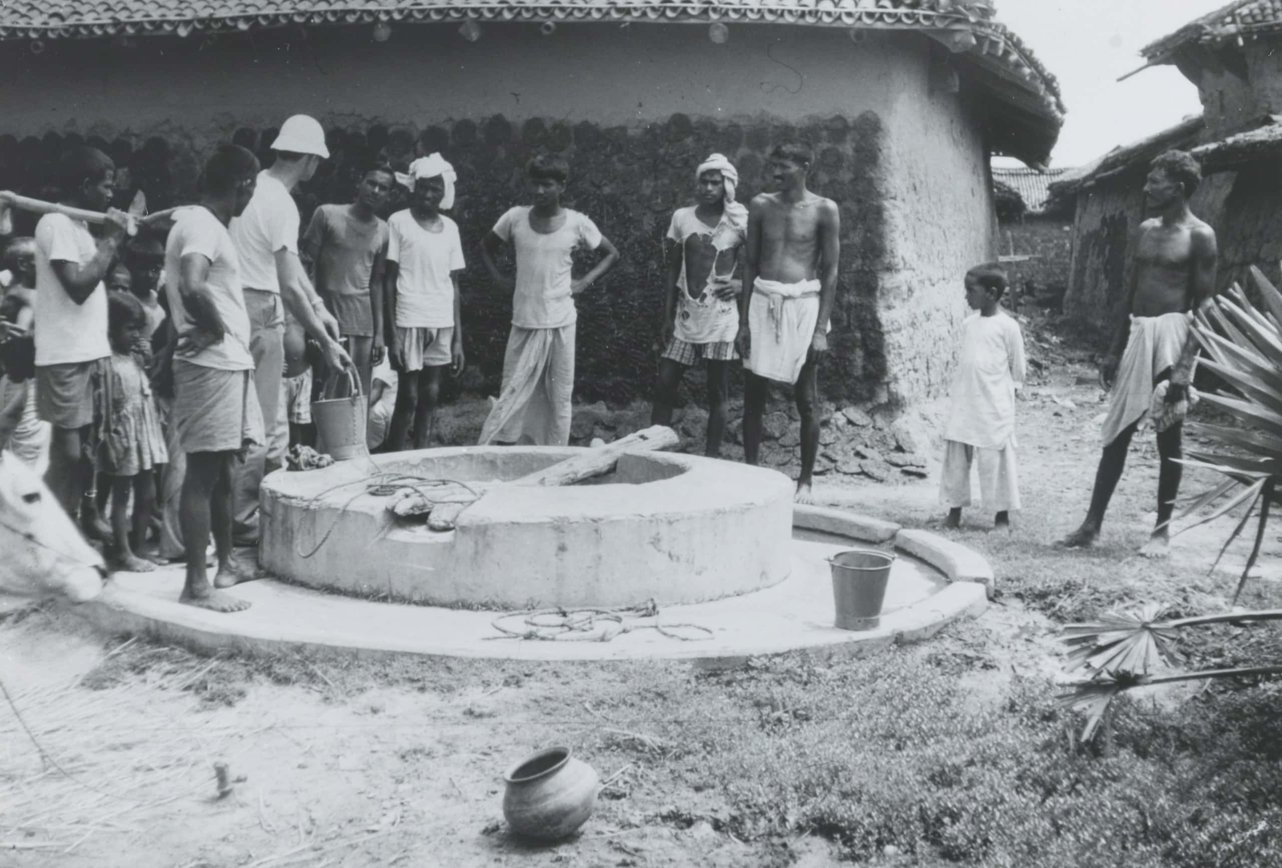 Zwart-witfoto, een groep mannen en kinderen staan om een waterput, velen van hen dragen versleten kleren. Op de grond staan emmers, op de achtergrond zijn lemen huizen te zien.