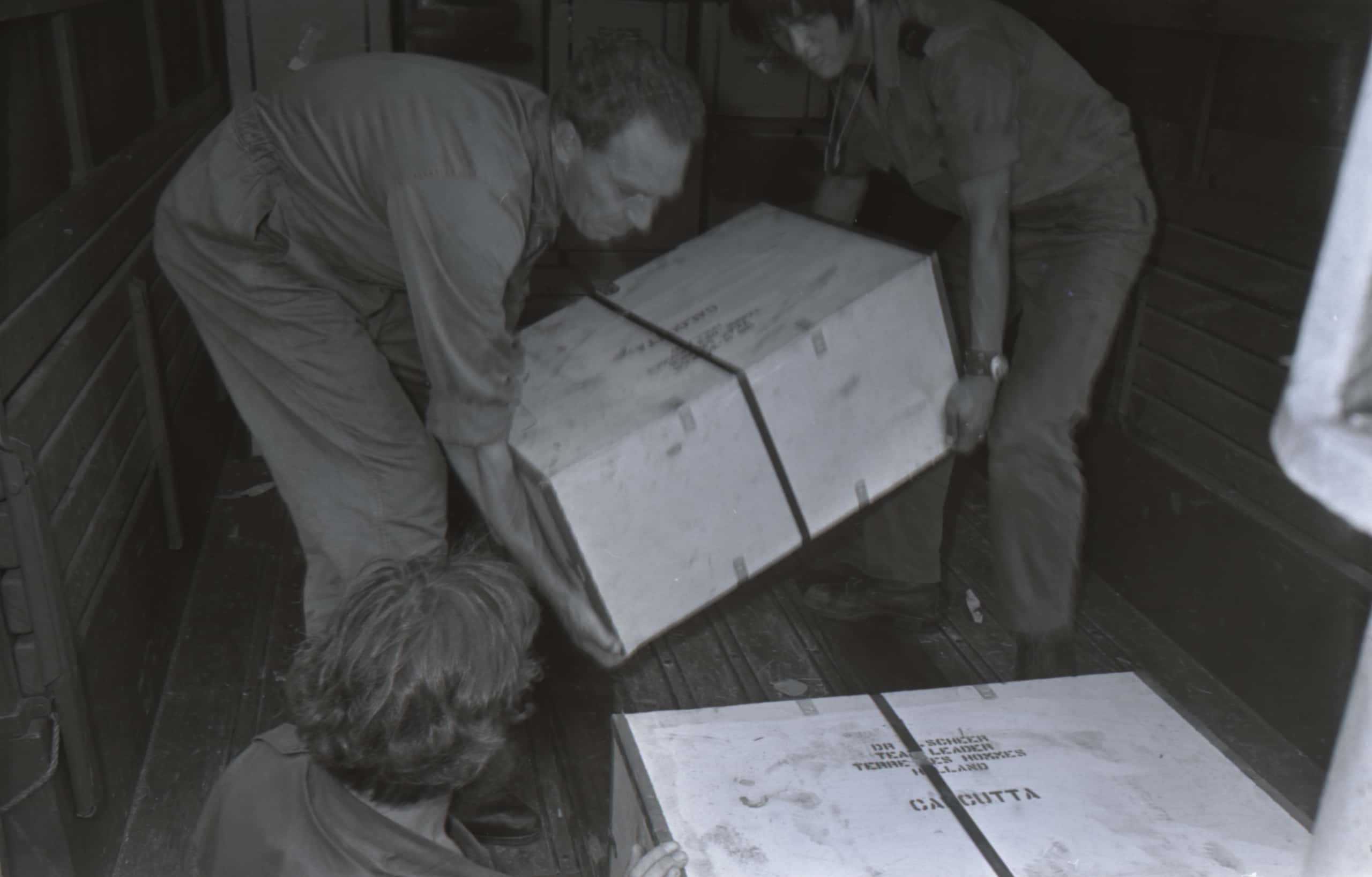 Zwart-witfoto. Twee militairen, één in overall en de ander in overhemd, zetten een houten kist op de grond van een transportvliegtuig. Om de kist zit een dunne, zwarte band.