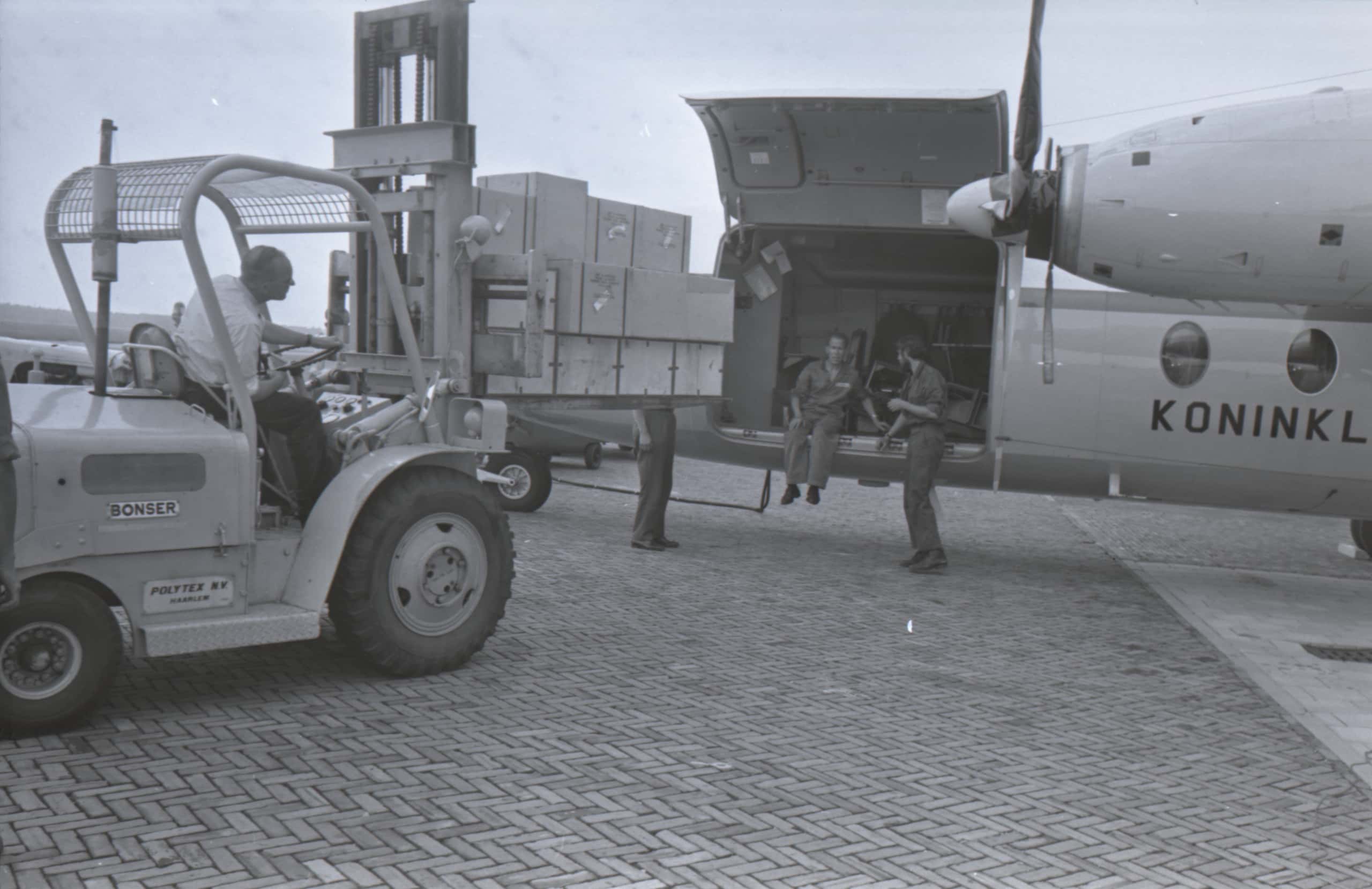 Zwart-witfoto. Een vorkheftruck rijdt naar een vliegtuig. Op de vorken staat een stapel van twaalf houten kisten. De zijdeur van het vliegtuig staat open, twee militairen zitten in de opening.