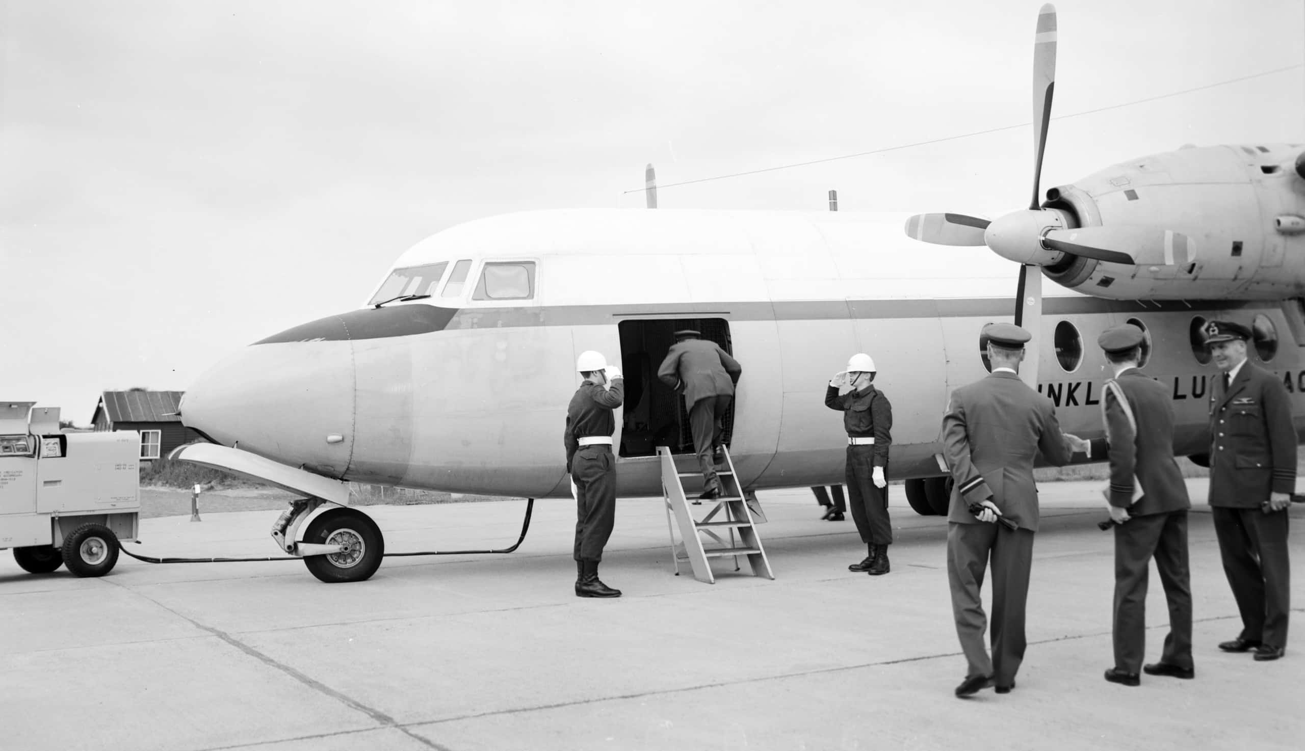 Zwart-witfoto. Te zien is een geparkeerd vliegtuig, in de romp zit een opening met een klein trappetje ervoor. Iemand stapt via de opening het vliegtuig binnen. Aan weerszijde van het trappetje staan twee militairen die groeten. Rechts op de voorgrond staan drie andere militairen die met elkaar in gesprek zijn, zij bewegen ook richting het vliegtuig. Links voor het vliegtuig is een tankwagentje voor de helft te zien, er loopt een slang vanuit het wagentje naar het vliegtuig.