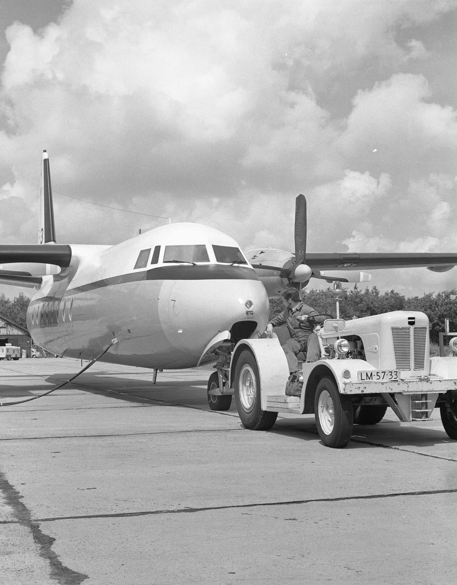 Zwart-witfoto. Een tractor trekt een vliegtuig voort. Op de tractor zit een man die omgedraaid naar achteren, naar het vliegtuig, kijkt. Op één van de vleugels is een motor met propeller te zien. In de achtergrond bomen en een hangar.