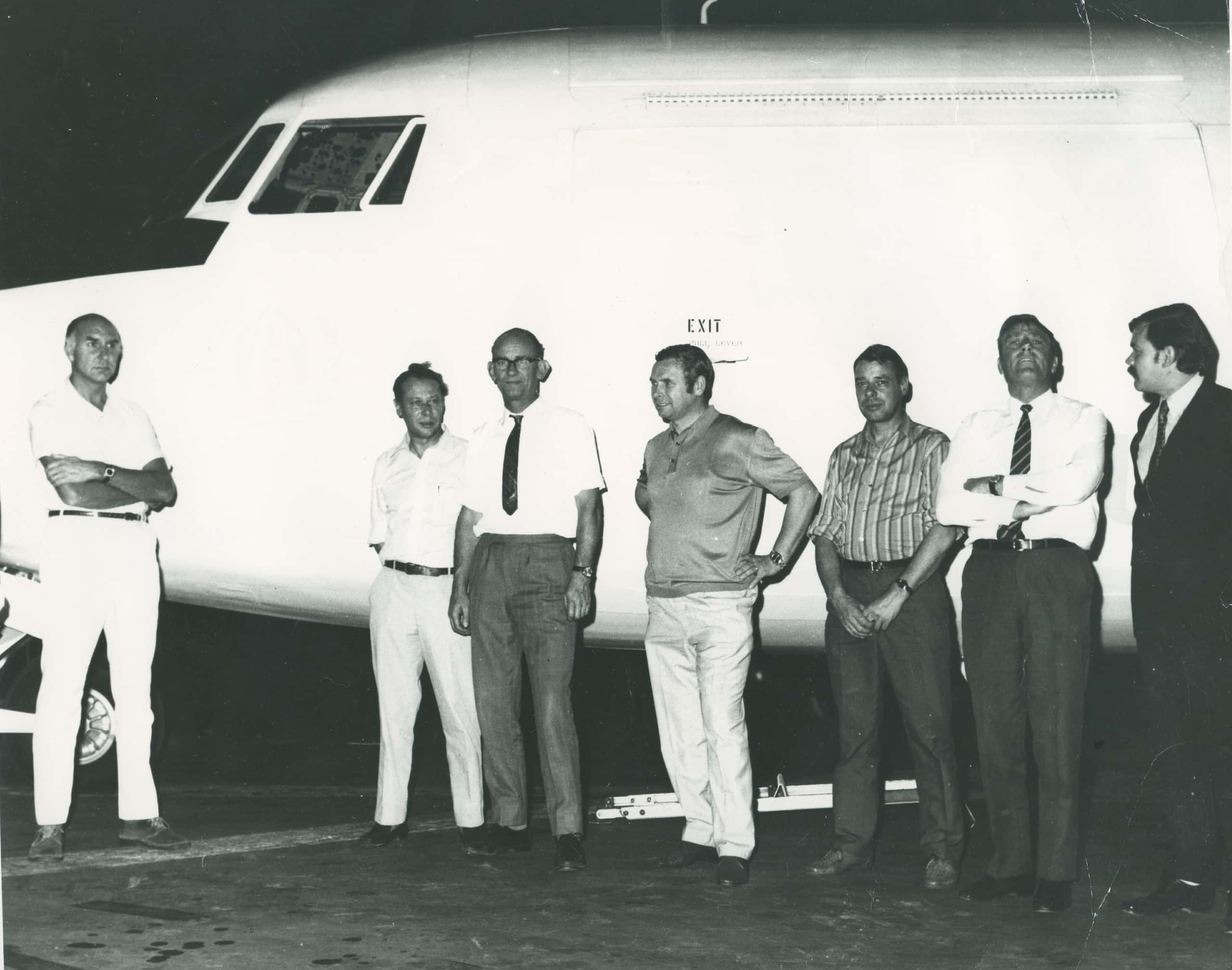 Zwart-witfoto, zeven mannen staan voor een wit vliegtuig, boven het hoofd van de middelste staat ‘exit’. De tweede van rechts en helemaal links staan met de armen over elkaar.