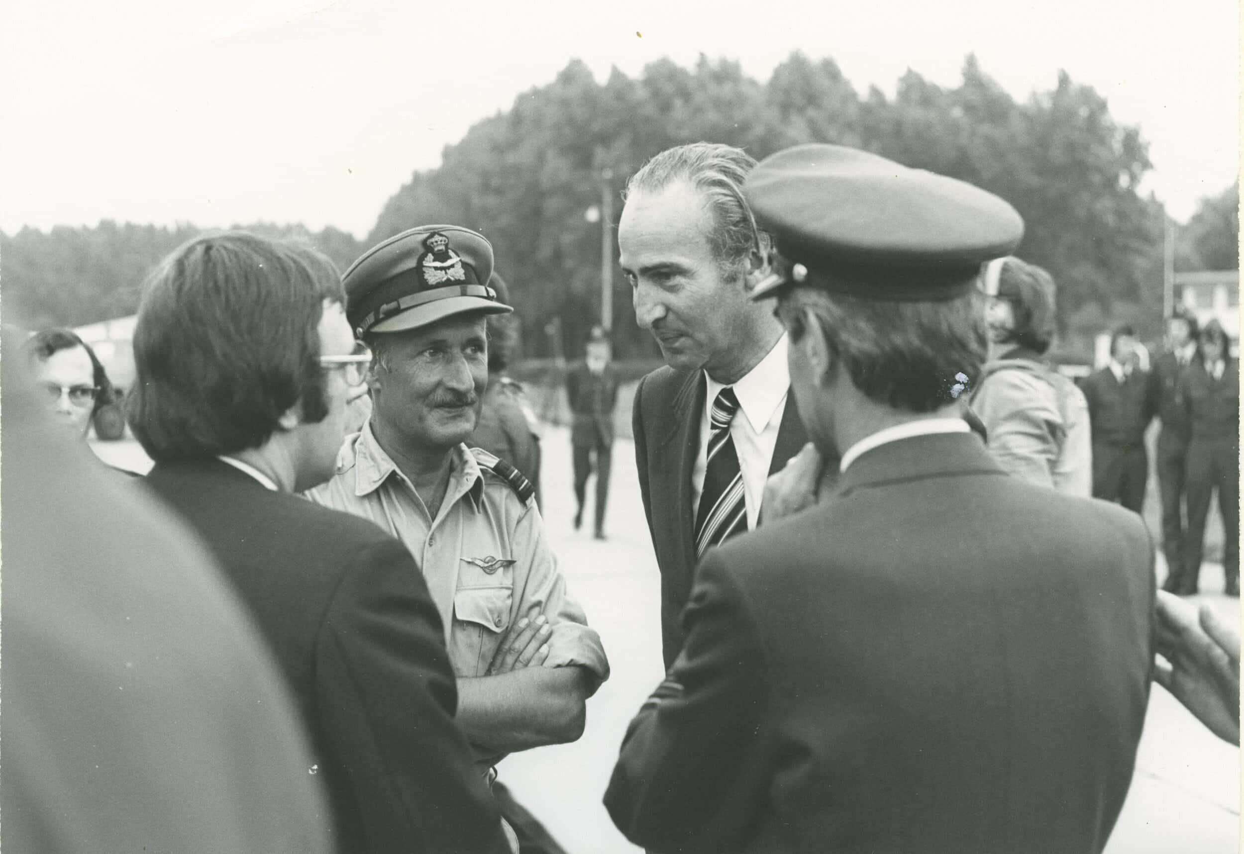 Zwart-witfoto, een groepje van vier mannen staan te praten, twee militairen, twee in pak, de twee militairen dragen een pet. Op de achtergrond meer militairen en journalisten.