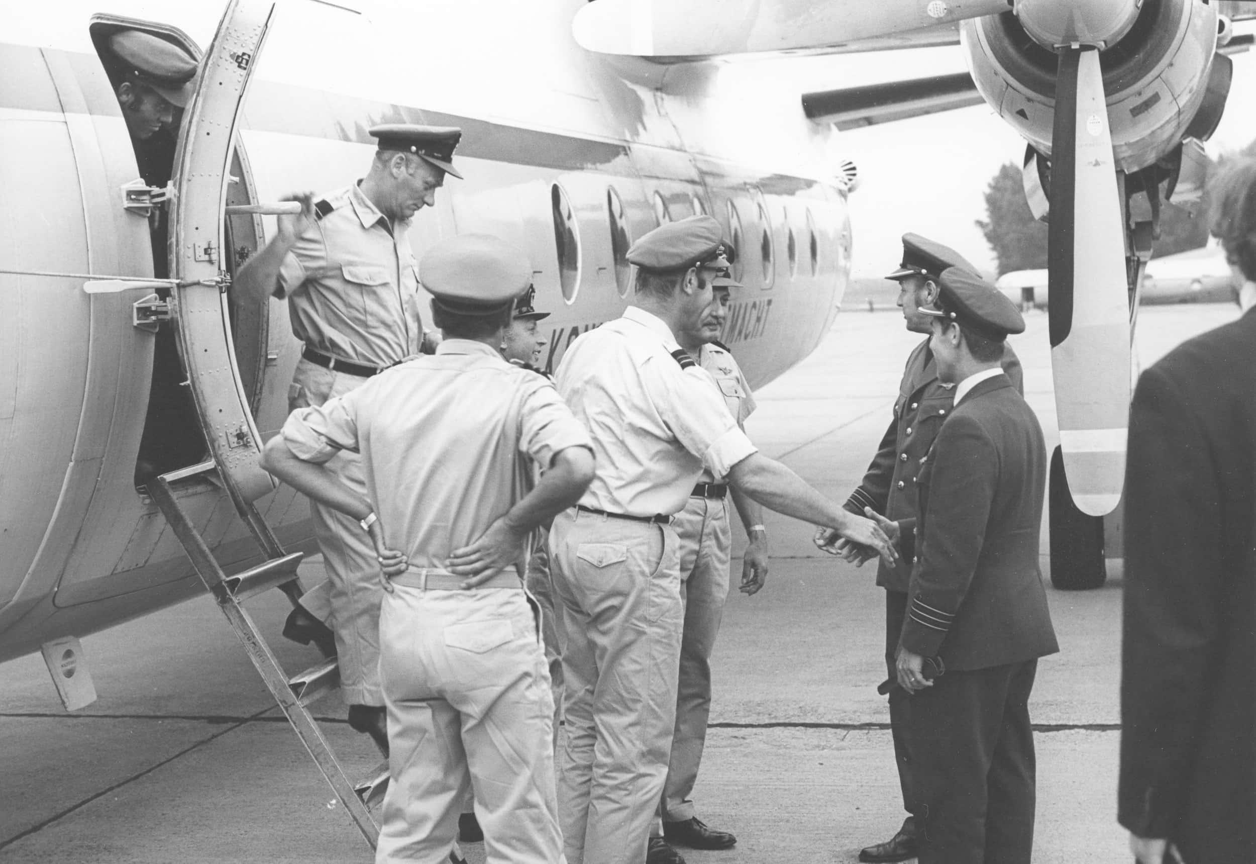 Zwart-witfoto, zes mannen staan naast een vliegtuig, één iemand stapt uit, een ander wacht op zijn beurt in de deuropening. Twee mannen op de grond schudden elkaar de hand.