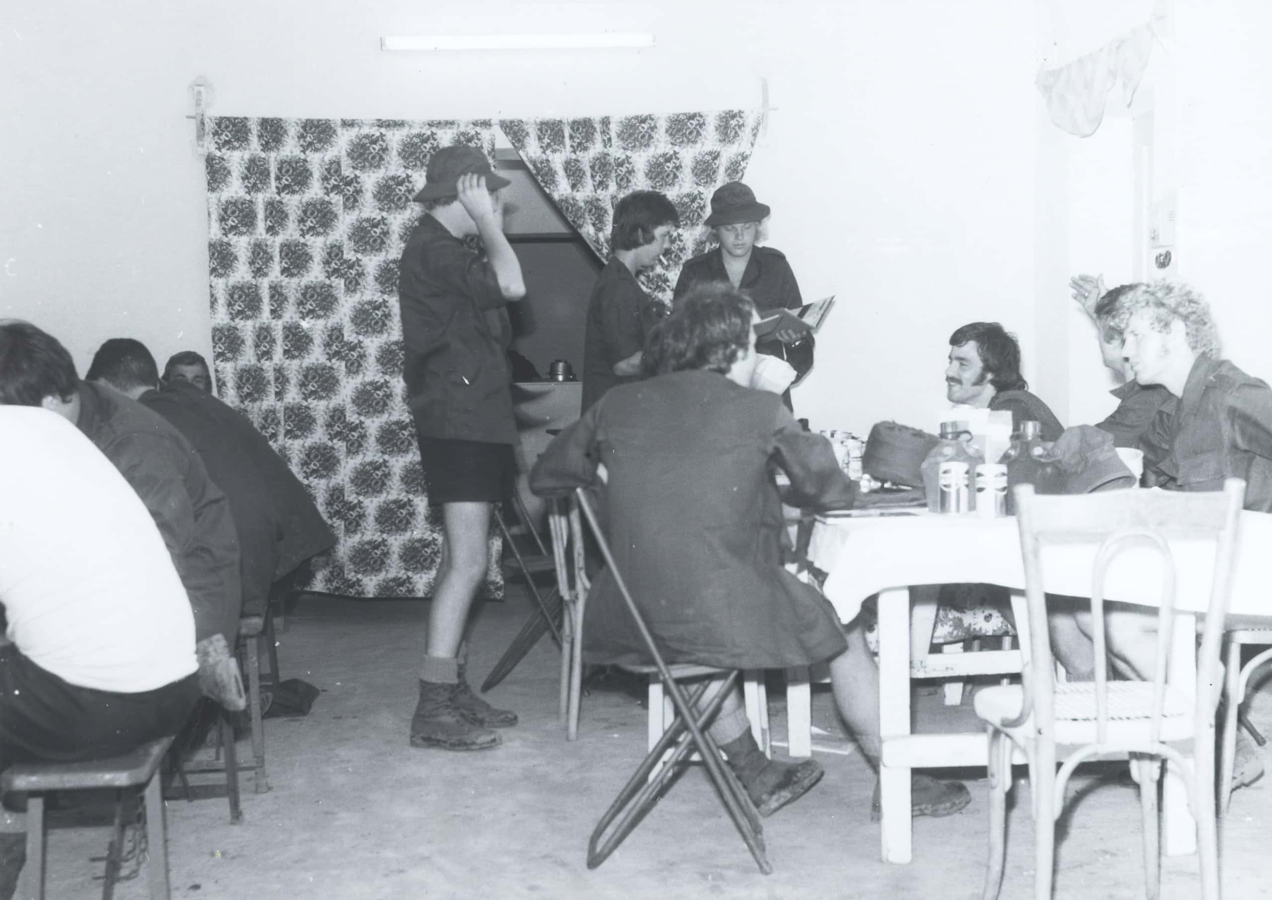 Zwart-witfoto, binnen. Militairen in een ruimte met tafels en stoelen, drie staan en de rest zit. Ze praten, lachen, drinken of lezen een tijdschrift. In het midden een militair die een zonnehoed opzet. Achter hem, midden in de muur, een opening waar twee grote bloemetjesgordijnen tot de grond voor hangen.