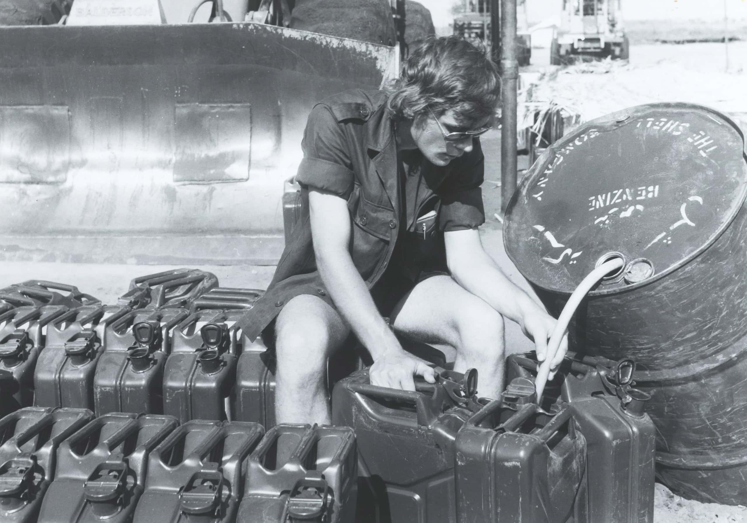 Zwart-witfoto, buiten. Een militair met zonnebril en in korte broek zit op twee jerrycans. Rechts naast hem een voorover hellende vat benzine, hier loopt een slangetje uit naar een jerrycans voor de voeten van de militair. Hij houdt met zijn linkerhand het slangetje vast en met zijn rechterhand een andere jerrycans. Overal om hem heen op de grond staan jerrycans in rijen naast elkaar. Links achter hem de bak van een wiellader.