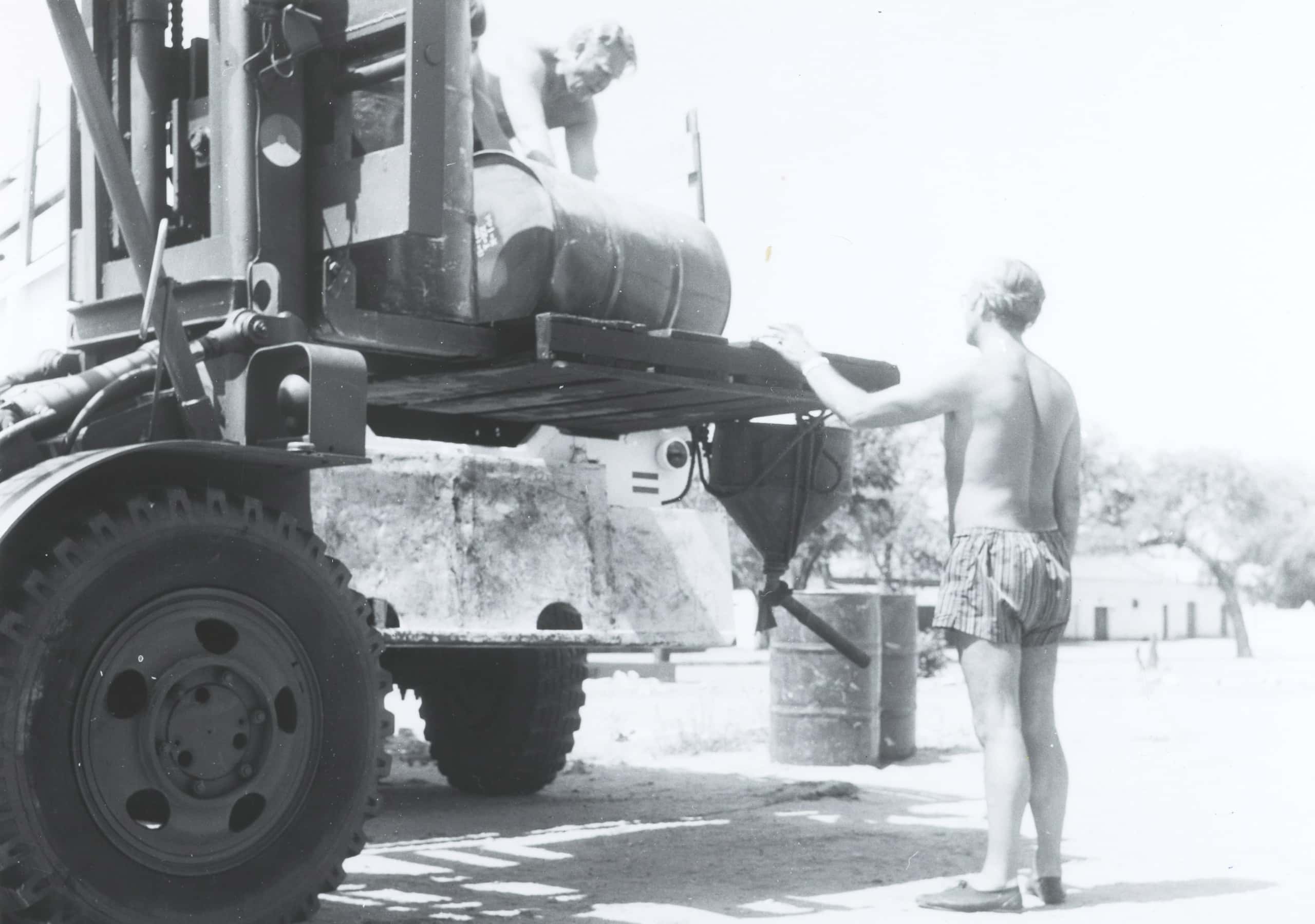 Zwart-witfoto, buiten. Een militair in alleen een korte broek staat naast een vorkheftruck, zijn linkerhand rust op het plateau van de truck voor hem, op schouderhoogte. Op het plateau zijn twee militairen bezig een benzinevat te rollen. Op de achtergrond twee benzinevaten op de grond, in de verte een gebouw en bomen.