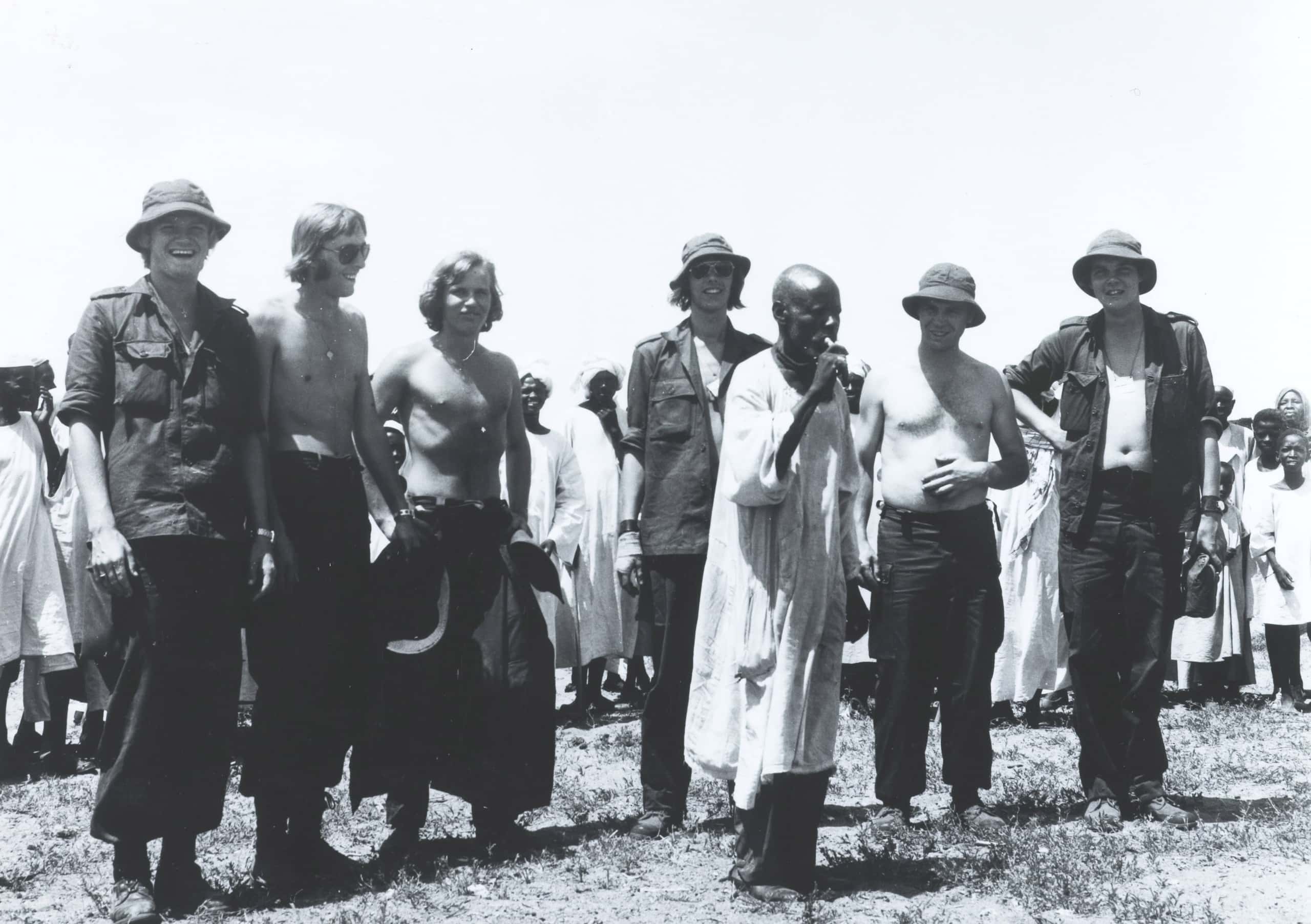 Zwart-witfoto, buiten. Een groep militairen staat op een rijtje naar elkaar, zij hebben hun shirts open of uit, sommigen dragen zonnebrillen en/of een zomerhoedjes. Zij lachen terwijl een kale man in lokale Afrikaanse kledij voor hen staan en op fluit blaast. Achter de militairen veel meer lokale mensen.