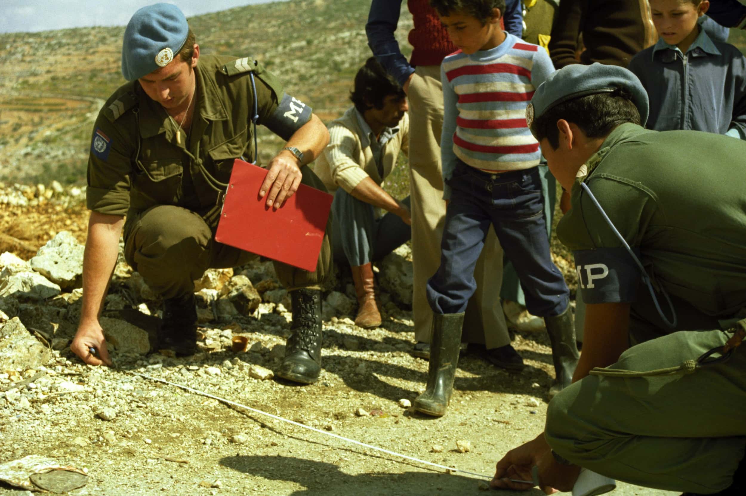 Kleurenfoto, buiten. Twee militairen zitten op hun hurken op een onverharde weg. De militair links heeft een clipbord in zijn ene hand en houdt met de andere hand een meetlint op de grond. De militair rechts houdt het andere eind van het meetlint vast. Naast hen staan meerdere kinderen mee te kijken.