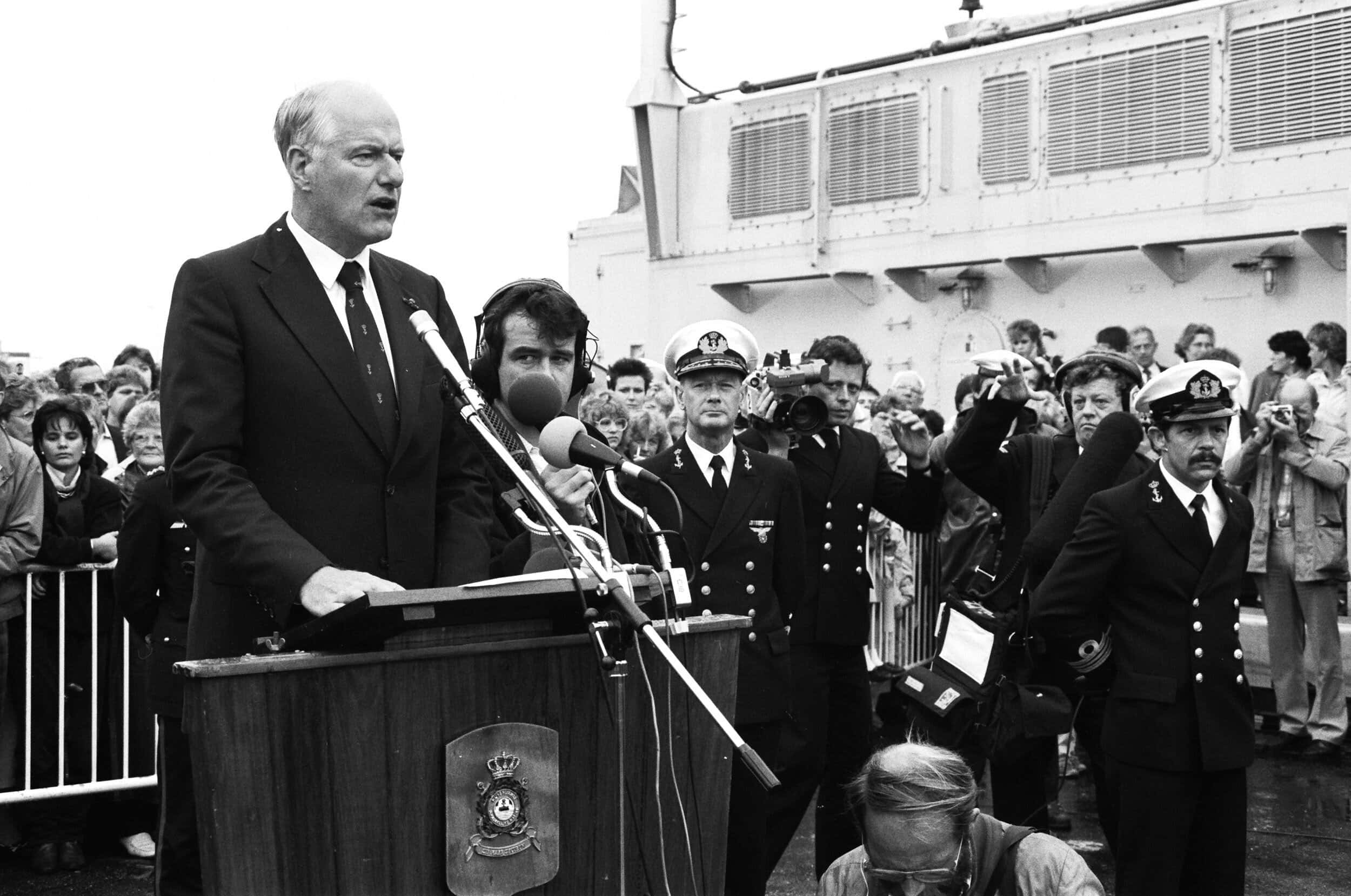 Zwart-witfoto, een man in pak houdt achter een katheter met drie microfoons een toespraak aan boord van een schip, rechts naast hem staan journalisten (sommigen met koptelefoon, camera of microfoon) en marineofficieren. Achter de spreker dranghekken met mensen erachter.