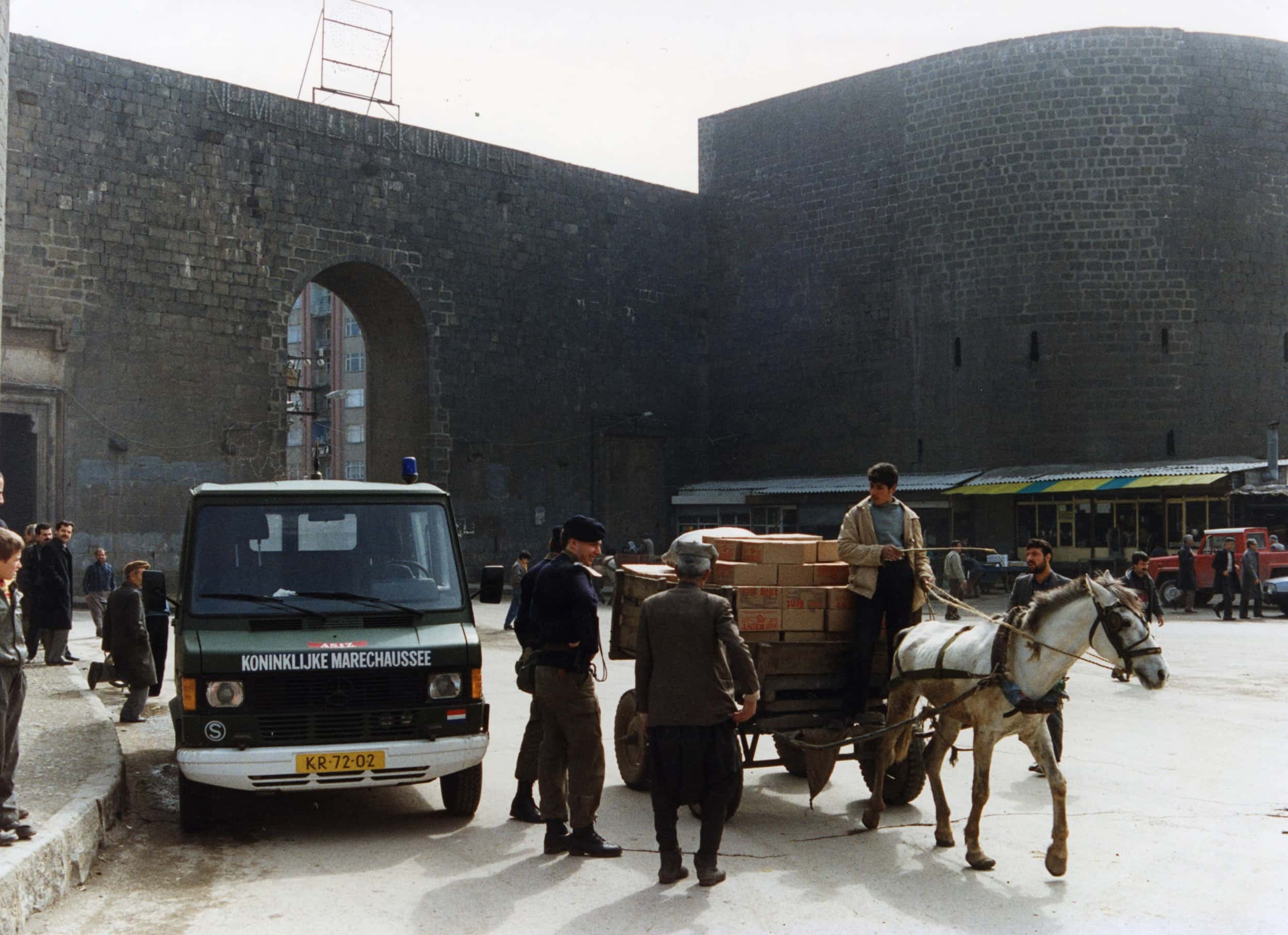 Kleurenfoto, straatbeeld van een stad. Twee militairen zijn op straat in gesprek met twee mannen in burger, één daarvan zit op een wagen volgeladen met dozen, dat wordt voortgetrokken door een paard. Links staat een groen busje met Nederlands kenteken tegen de stoep geparkeerd, daaromheen staan meerdere mensen. Op de achtergrond een hoge bakstenen stadsmuur met links van het midden een hoge poort.