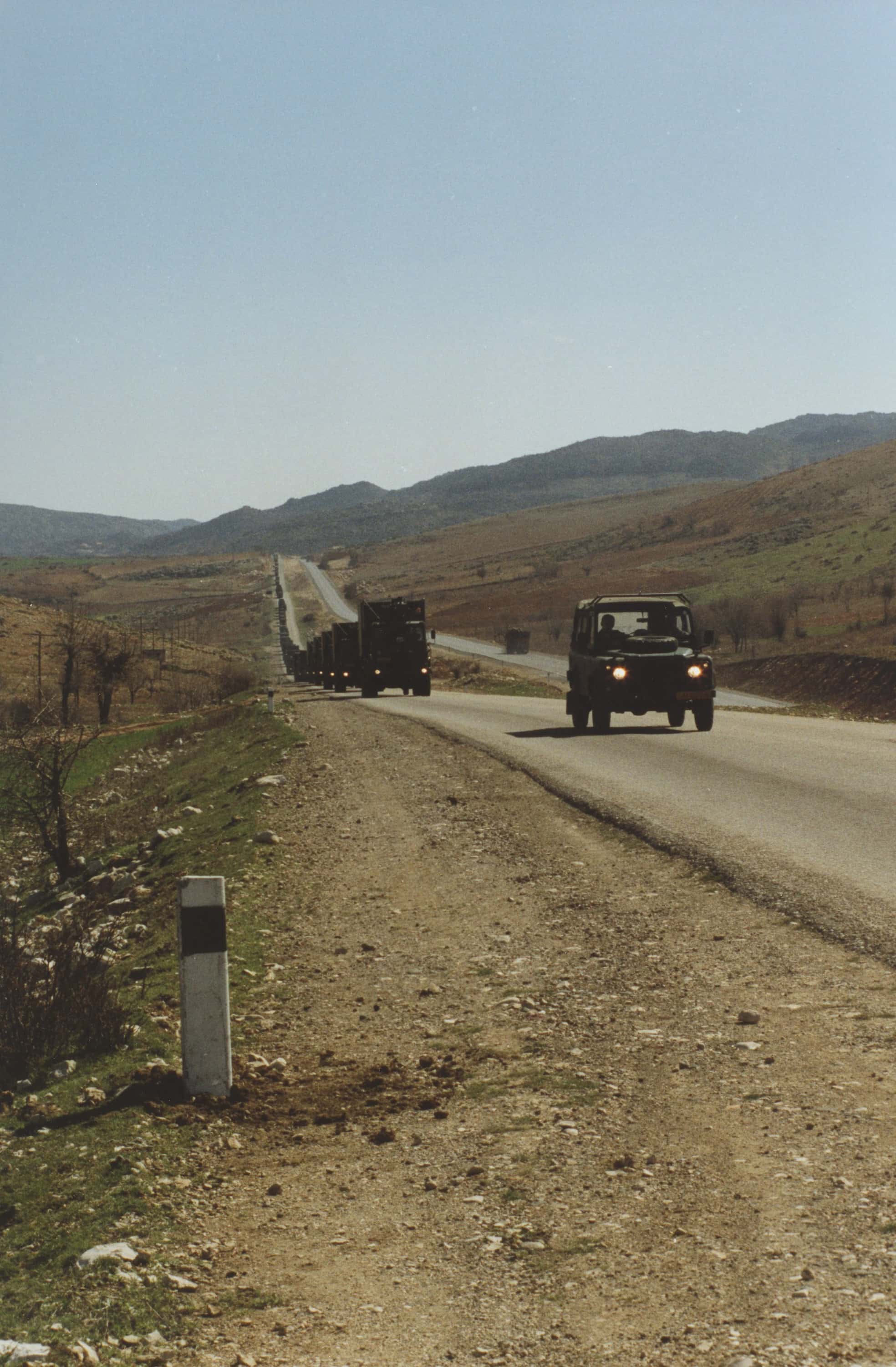 Kleurenfoto, buiten langs de kant van de weg. Over een geasfalteerde weg komt een lange rij vrachtwagens aanrijden die aan de horizon uit beeld verdwijnt, daarvoor rijdt een jeep. Het landschap is droog, dor een bergachtig, met soms gras en hier en daar een boom of lage struik.