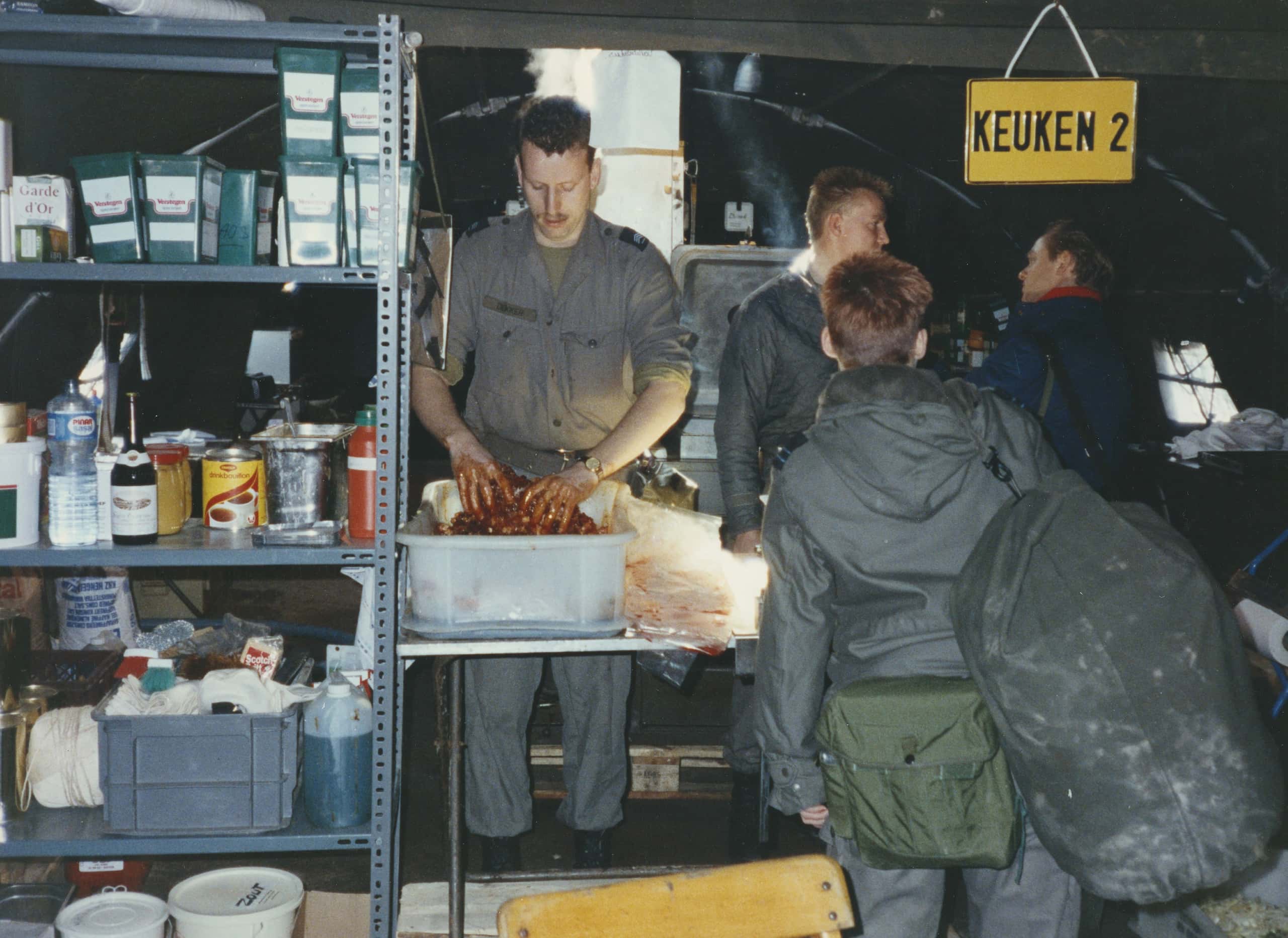 Kleurenfoto, binnenkant van een militaire tent. In het midden een man met snor in legergroen overall die met zijn handen in een plastic bak eten aan het bereiden is, zijn handen zijn bedekt met een roodbruine saus. Rechts een vrouw op de rug gezien, met een plunjebaal over haar rechterschouder. Boven haar hoofd hangt een bordje met de tekst Keuken 2. Links een stellingkast met vier planken met allerlei pakken en bakken aan eet- en drinkwaren.