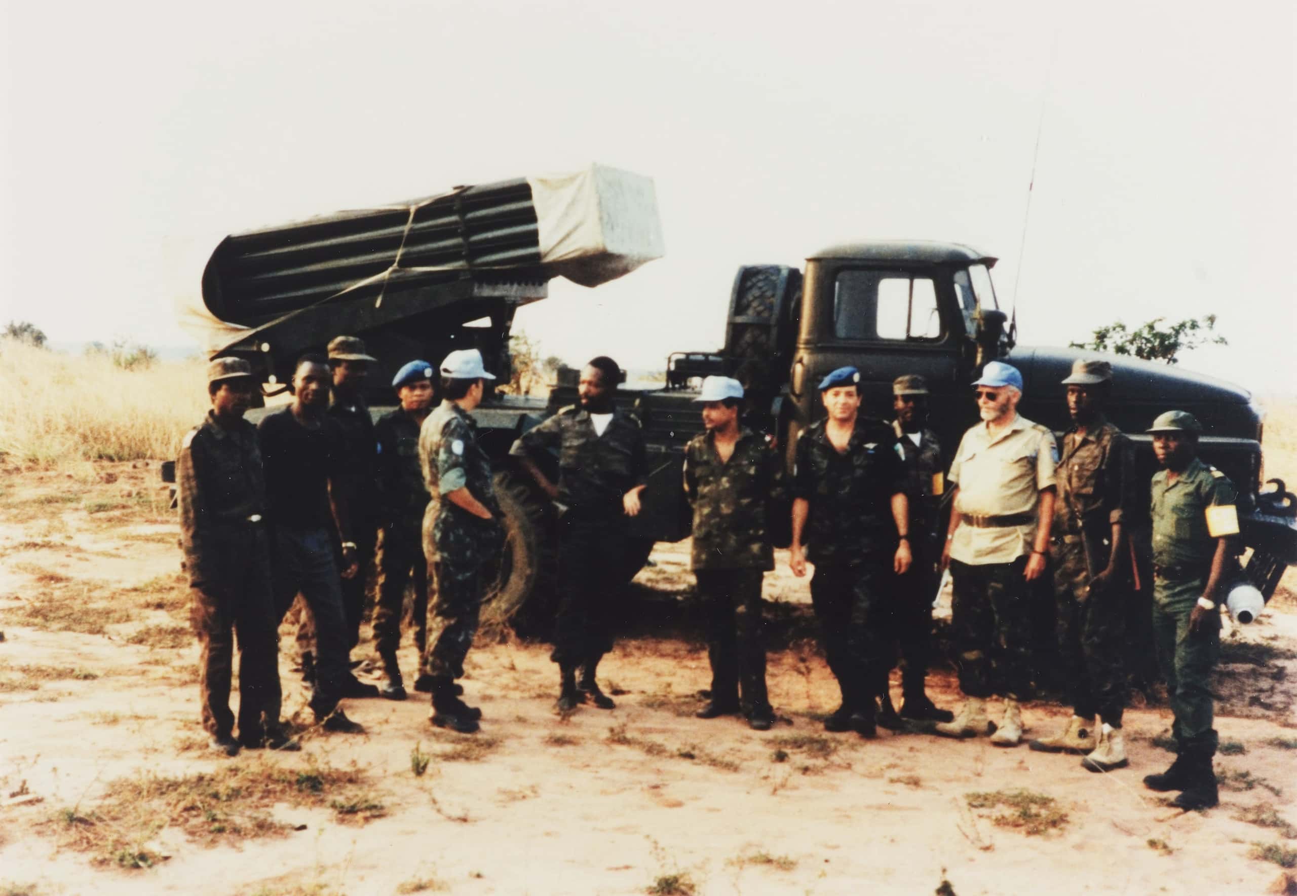 Kleurenfoto, buiten. Een groep van twaalf militairen staat voor een vrachtwagen met een raketlanceringssysteem. Ze dragen allemaal camouflagekleding, vier van hen dragen een blauwe baret of pet. Sommigen kijken in de camera, anderen zijn met elkaar in gesprek. Het landschap is zanderig en dor, met wat gras.