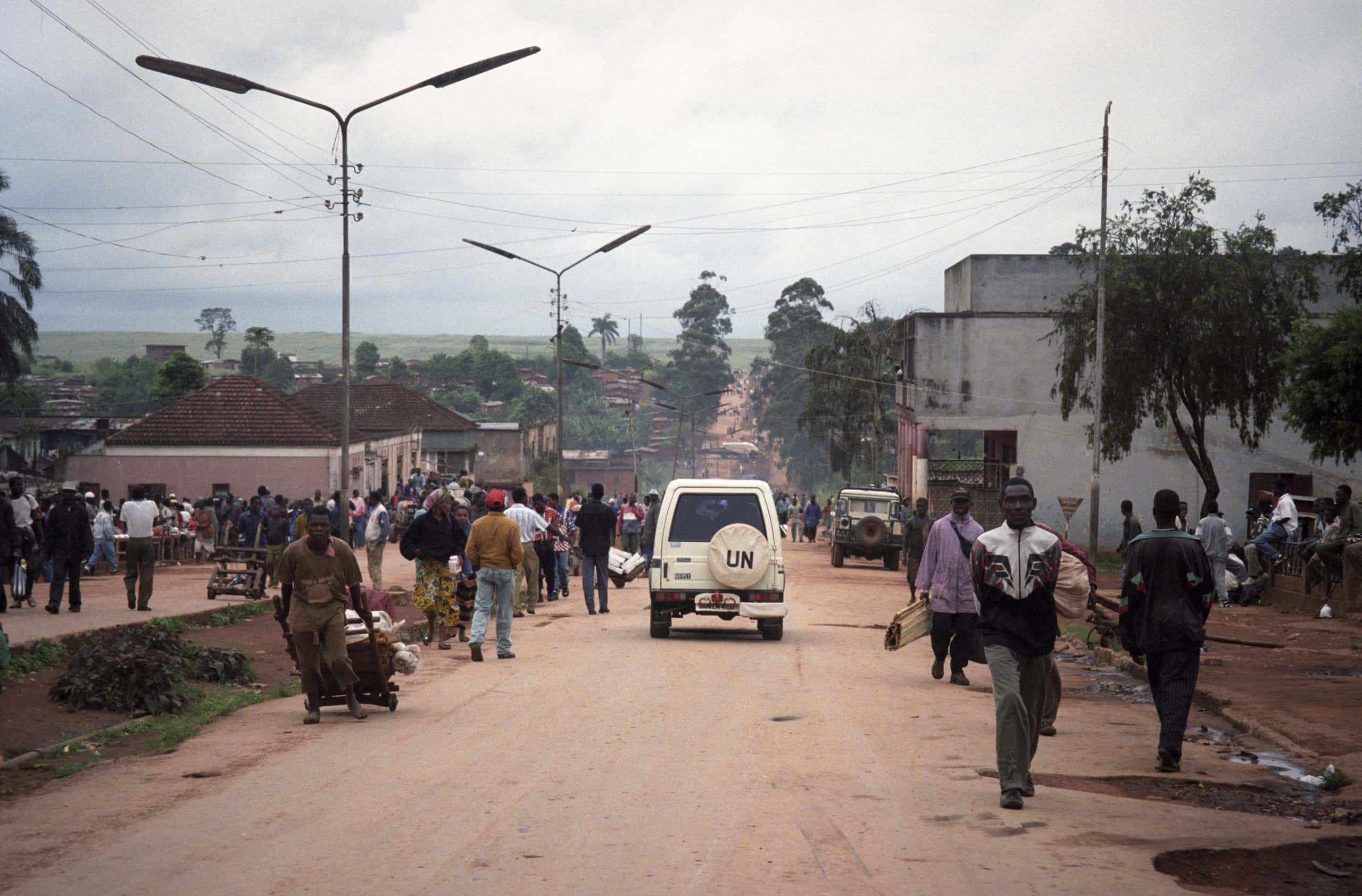 Kleurenfoto, buiten op straat. Straatbeeld van een Afrikaanse stad, centraal op de foto rijdt een witte auto op de onverharde weg, met achterop de letter UN op de reserveband. Er lopen veel mensen op straat en op de stoep, op de achtergrond huizen en bomen, aan de lantaarnpalen zijn meerdere elektriciteitsdraden gevestigd.