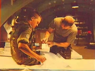 Kleurenfoto, binnen. Een militair in camouflagekleding zit op een bed of tafel, hij houdt zijn linkerhand voor zich. Rechts een militair die voorovergebogen de uitgestoken hand vasthoudt en bekijkt. Op de hand ligt een wit doekje of verband.