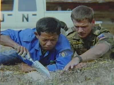 Kleurenfoto, buiten in een grasveld. Twee mannen liggen in het gras, de linker draagt een blauwe overall, de rechter draagt camouflagekleding. De linker heeft een troffel in zijn hand en staat op het punt om te graven, terwijl de rechter aanwijst waar dat moet gebeuren. Op de achtergrond een witte truck met de letters UN erop.