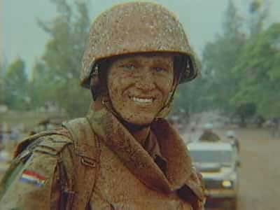 Kleurenfoto, buiten. Een militair in camouflagekleding en helm kijkt in de camera en lacht. Zijn kleding, gezicht en helm zitten bijna helemaal onder de modder. Alleen zijn tanden en het Nederlandse vlaggetje op zijn mouw niet. Op de achtergrond een witte truck met twee militairen achterop en daarachter bomen en struiken.